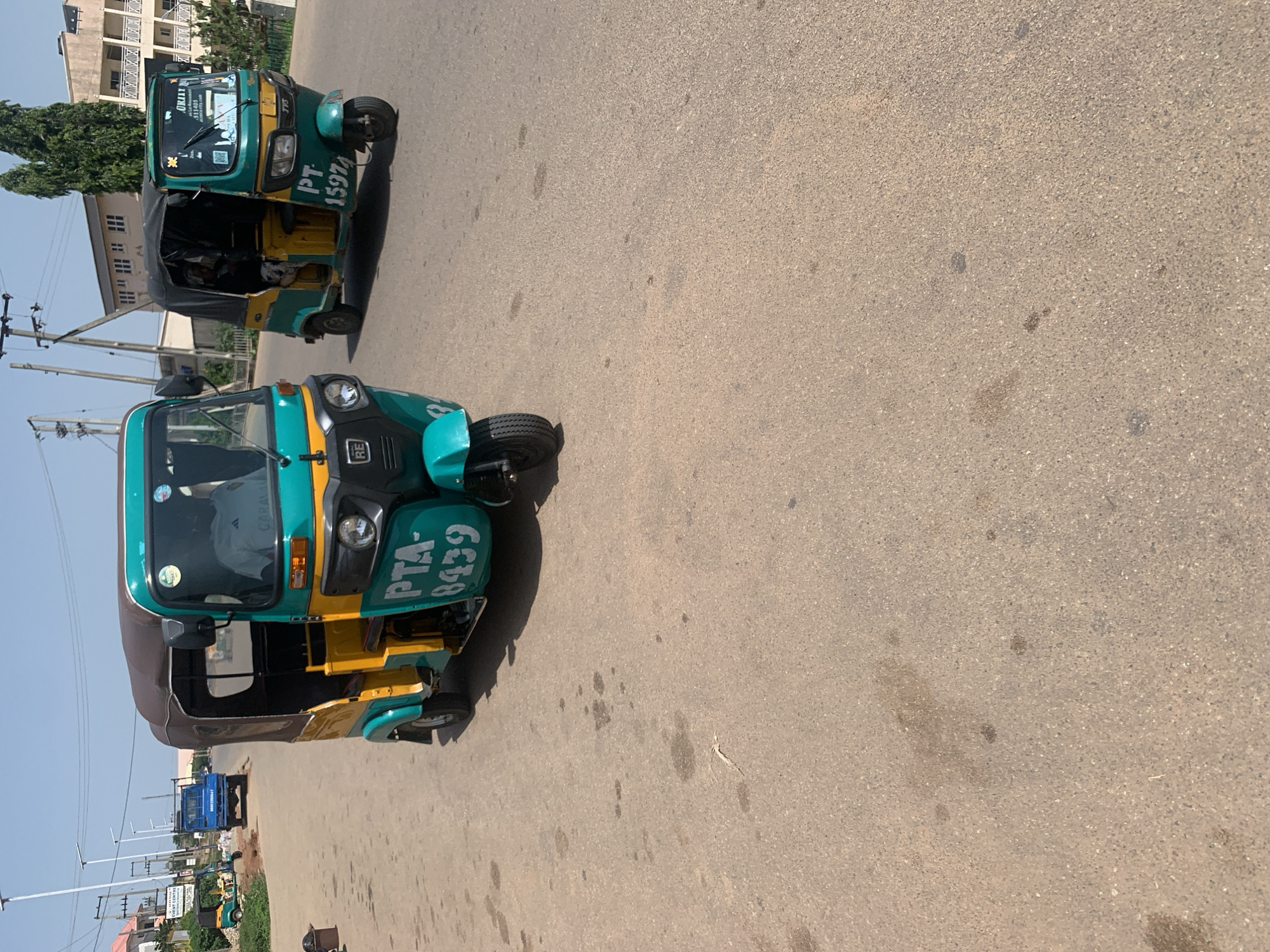 The image depicts a street scene in Jos, Nigeria, featuring two auto-rickshaws, commonly known as "Keke Napep." The auto-rickshaws are painted in a distinctive green and yellow color scheme. The foreground auto-rickshaw has the registration number "PTA-8439" clearly visible on its side. The second auto-rickshaw, positioned behind and slightly to the left, shows the registration "PT 15974." Both vehicles appear to be parked or waiting on a paved road under a clear, sunny sky, suggesting daytime.
In the background, several buildings with a modern architectural style are visible, along with trees and streetlights. The presence of these elements indicates an urban or semi-urban environment. There are no people actively driving or riding in the auto-rickshaws, suggesting they might be parked or waiting for passengers. The road surface shows some dark stains, possibly oil or water marks. The overall scene conveys a typical day in a Nigerian city, with common modes of transportation evident. The lighting suggests it is likely mid-morning or early afternoon.