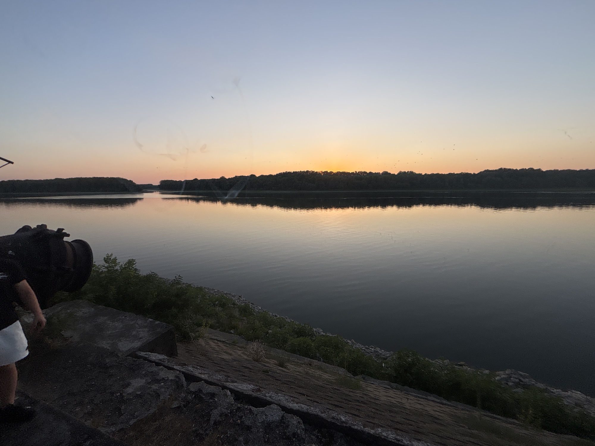 The image depicts a serene sunset scene over a calm body of water, likely a river or a lake, with a tree-lined horizon. The sky transitions from a pale blue at the top to warm hues of orange and yellow near the horizon, indicating the sun is setting or has just set. Several small dark specks, possibly birds, are visible in the sky. In the foreground, on the left side, a person wearing a black t-shirt and white shorts is partially visible, standing near a large, dark, industrial-looking object with a pipe-like protrusion. The ground in the foreground is made up of rough concrete and rocks, with some vegetation growing along the edge of the water. The water itself is dark and reflects the colors of the sky. The overall impression is one of natural beauty and tranquility, with a hint of industrial presence due to the object in the foreground. The location is identified as Marten, Bulgaria.