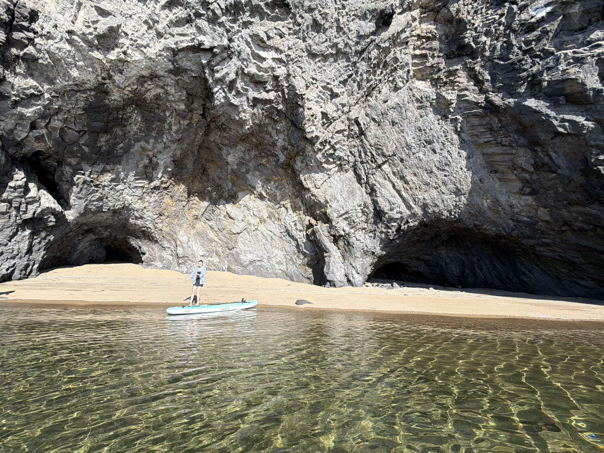 The image shows a person standing on a paddleboard in clear, shallow water. The paddleboard is light blue and has the brand name "JALE DI" and "10'6" visible. The person is wearing a blue long-sleeved shirt and black shorts. They are facing away from the camera, looking towards a sandy beach and a large, rocky cliff face. The cliff has several cave-like openings. The water is calm with sunlight reflecting off the ripples, creating a dappled effect. The sand is light brown and appears dry. The sky is bright blue with no clouds visible. The overall scene suggests a sunny day at a natural coastal location. The location is identified as an unknown city in Russia.