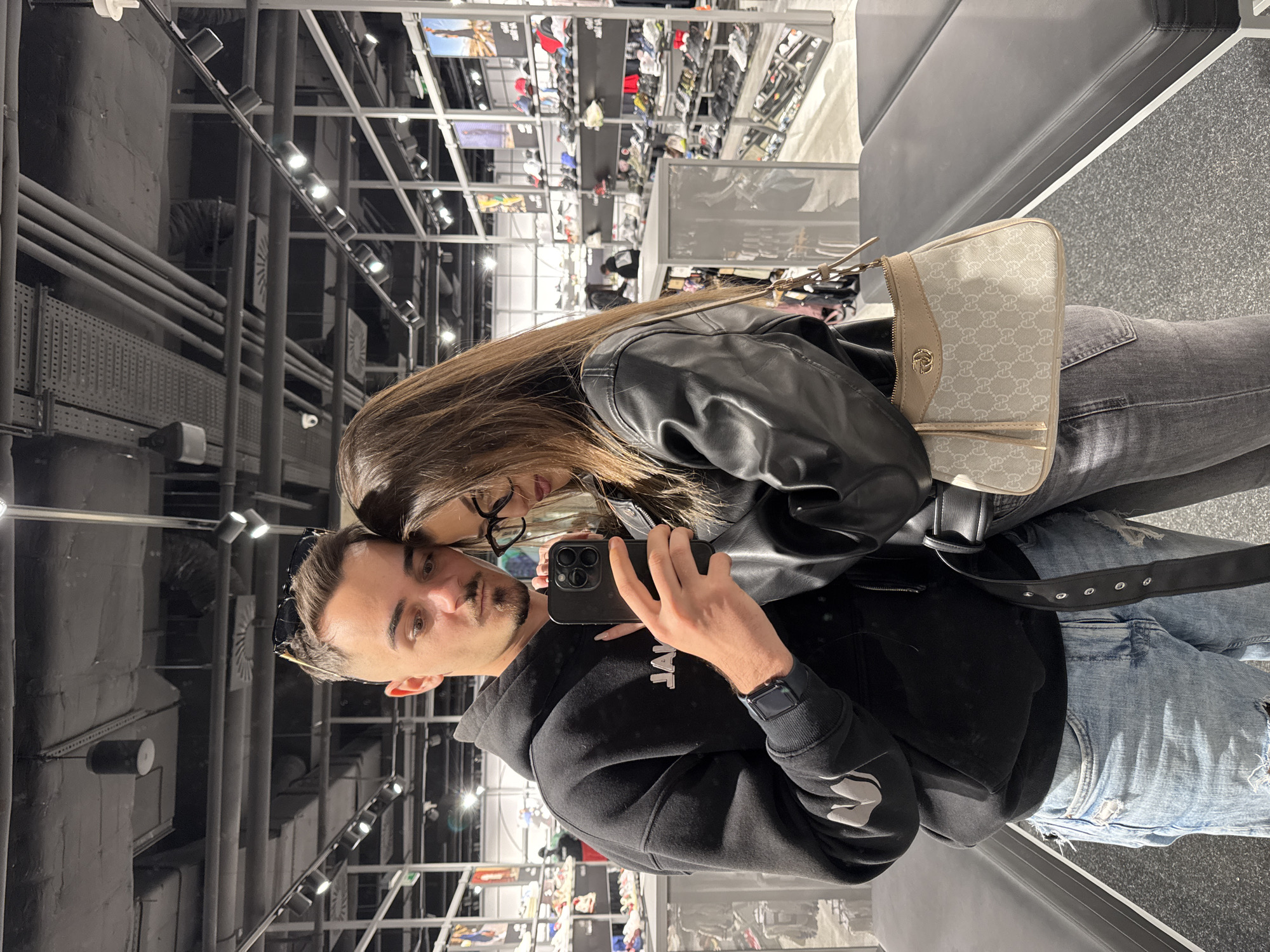 A couple is taking a selfie in what appears to be a retail store in Sofia, Bulgaria. The man is wearing a black hoodie and distressed jeans, holding a smartphone. The woman is in a black leather jacket and grey jeans, leaning into him. A patterned Gucci bag is visible. The background shows a store environment with rows of shelves stocked with merchandise, presumably footwear. The store's ceiling structure is exposed, with various lighting fixtures and ventilation systems visible.