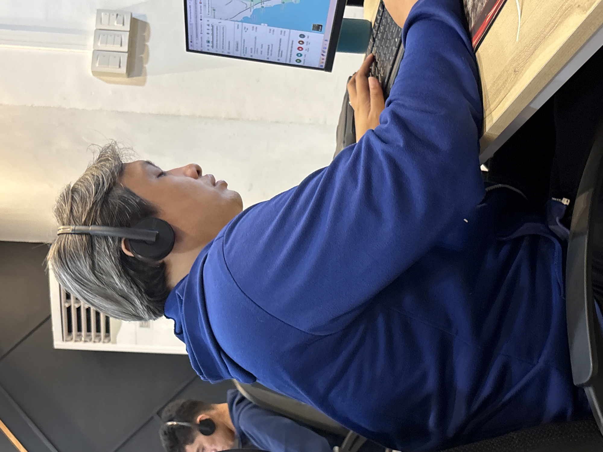 The image shows a young man wearing a blue hoodie and headphones, sitting at a desk and looking at a computer screen. He is in a room with white walls and a wooden desk. There is another person in the background, also wearing headphones. The computer screen shows a map with some data points. The scene appears to be an office or a call center. The location is described as being in Davao City, Philippines.
