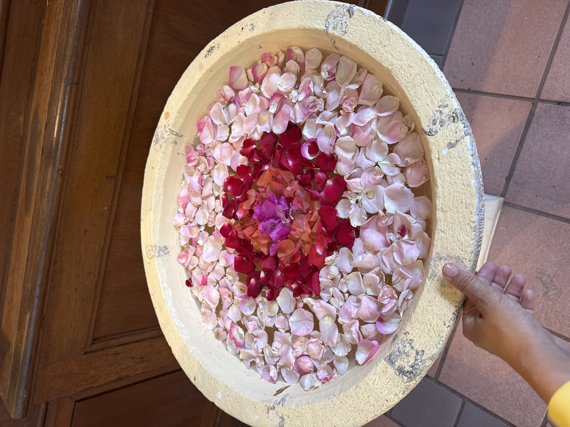 The image shows a top-down view of a shallow, round, beige-colored bowl filled with water and flower petals. The petals are primarily light pink and white, with a cluster of dark red and purple petals concentrated in the center. The bowl sits on a surface with terracotta-colored tiles, and a portion of a wooden door is visible on the left. A human hand with tan skin is touching the rim of the bowl on the right side. The overall scene is located in Kuta Selatan, Indonesia.