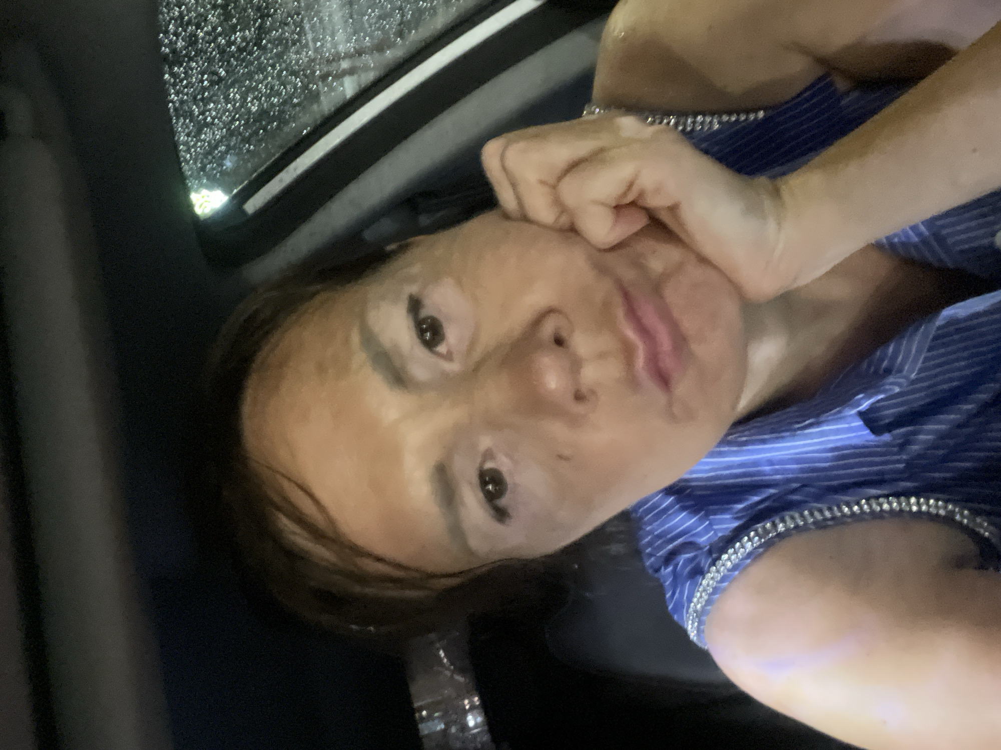 A selfie of a woman in a car at night. The woman has brown hair and is resting her chin on her hand. She is wearing a blue striped shirt with rhinestones on the straps. The car window is visible on the right side of the image, with raindrops on it. The background is dark, with some out-of-focus lights visible. The overall impression is a candid, slightly melancholic moment.
