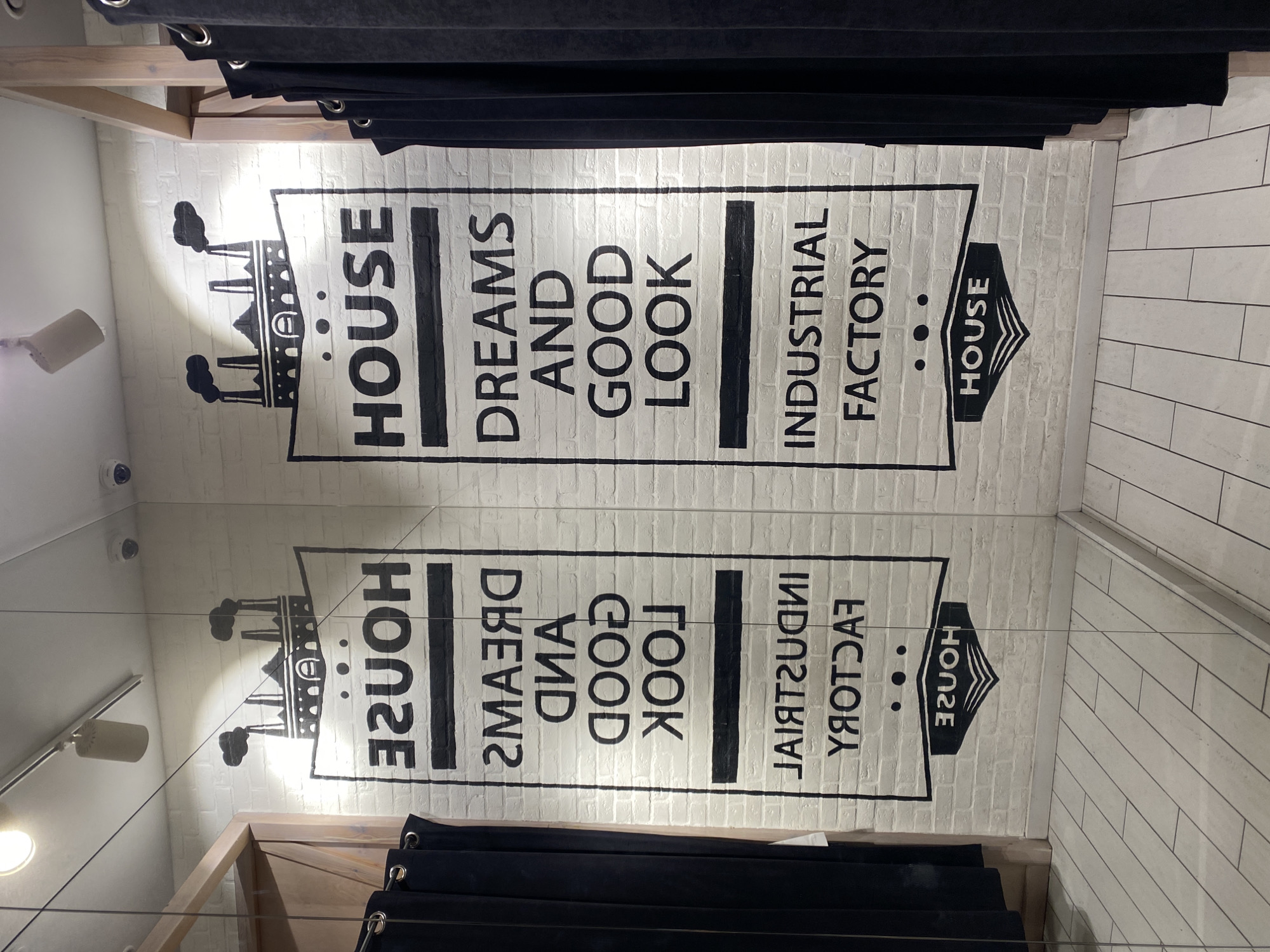 The image captures the interior of a fitting room or dressing room in a clothing store. The decor features a wall mural in black lettering and illustrative elements on a white, brick-like background, which appears to be mirrored on a wall, creating a symmetrical effect. The mural includes stylized factory imagery with smokestacks, the word "HOUSE" in a large font, followed by stacked phrases "DREAMS AND GOOD LOOK" and "INDUSTRIAL FACTORY". The bottom right of the mural concludes with a logo in a stylized arrowhead shape that also includes the word “HOUSE”.
Black curtains are suspended from a track, obscuring the top and bottom portions of the image, adding a sense of enclosure. Additionally, visible hardware, such as curtain rings and a wooden frame supporting the curtain rail, are present at the top of the image. On the left, lighting fixtures or security cameras are mounted on the wall, as the reflection in the mirror displays additional lighting. The background shows part of a wall with a simple, uniform design, while other walls seem to be mirrored as well.