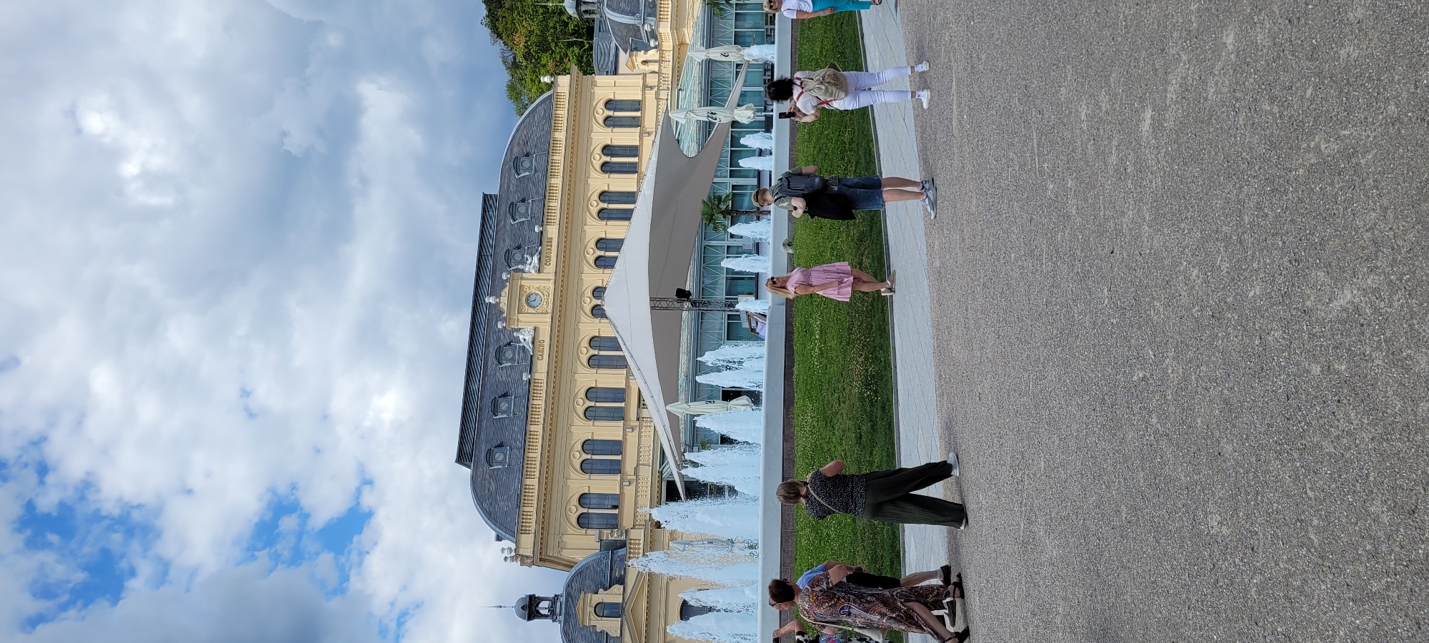In the city of Baden, Austria, a group of people are visible in front of the grand Casino Baden building, a large, yellow structure with a dark roof and numerous arched windows. A row of water fountains sprays upwards in front of the building, with a large white canopy partially obscuring the view. The ground is paved with grey gravel. Several individuals are walking or posing for photos. One woman in a pink dress stands by herself, looking towards the camera. Another woman in a patterned dress walks away from the viewer, while a man in a black shirt and dark shorts stands beside her. Further to the right, a man with a backpack is captured in profile, and a woman with a white backpack is taking a photo with her phone. The sky above is partly cloudy.