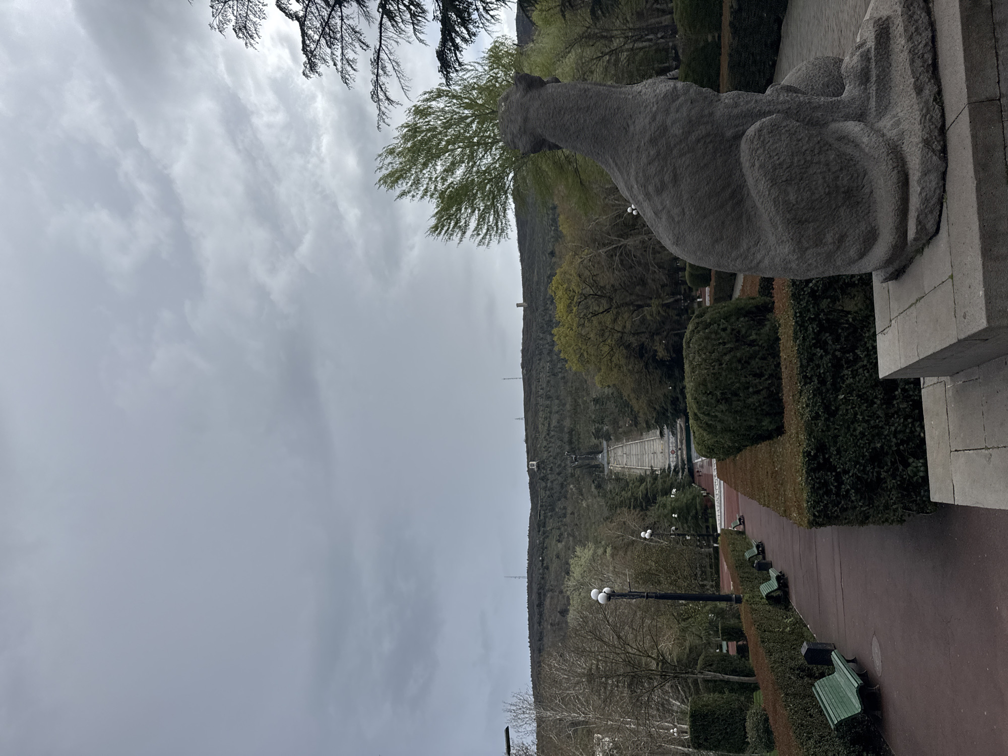 This image depicts a park in Tbilisi, Georgia, featuring a prominent stone sculpture of a reclining lion or panther-like creature overlooking a broad pathway leading toward a mountain. The sky is overcast with heavy clouds. The sculpture sits atop a stone platform or pedestal. A paved walkway extends into the distance, flanked by neatly trimmed hedges and rows of benches. Lampposts line the walkway. In the background, a hill rises steeply, partly covered with trees and featuring a distant monument or statue complex at its summit. The general atmosphere suggests a cool, possibly overcast day in a well-maintained urban park.