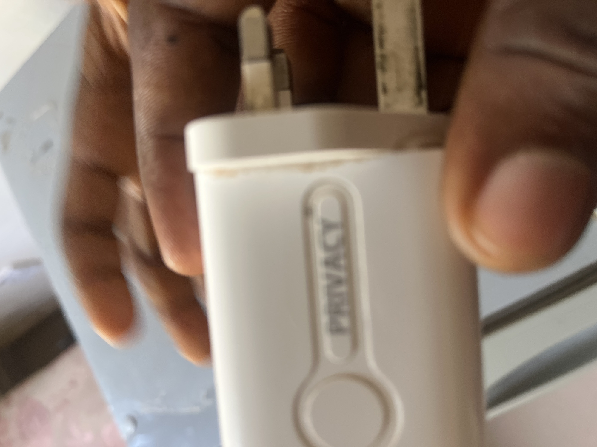 A person's hand is holding a white electrical adapter. The adapter has the word "PRIVACY" embossed on it, along with a circular indentation to its left. The adapter has three metal prongs extending from its right side. The background is blurred, showing what appears to be a light-colored surface. There is no information in the image to suggest a specific location in Jalingo, Nigeria.