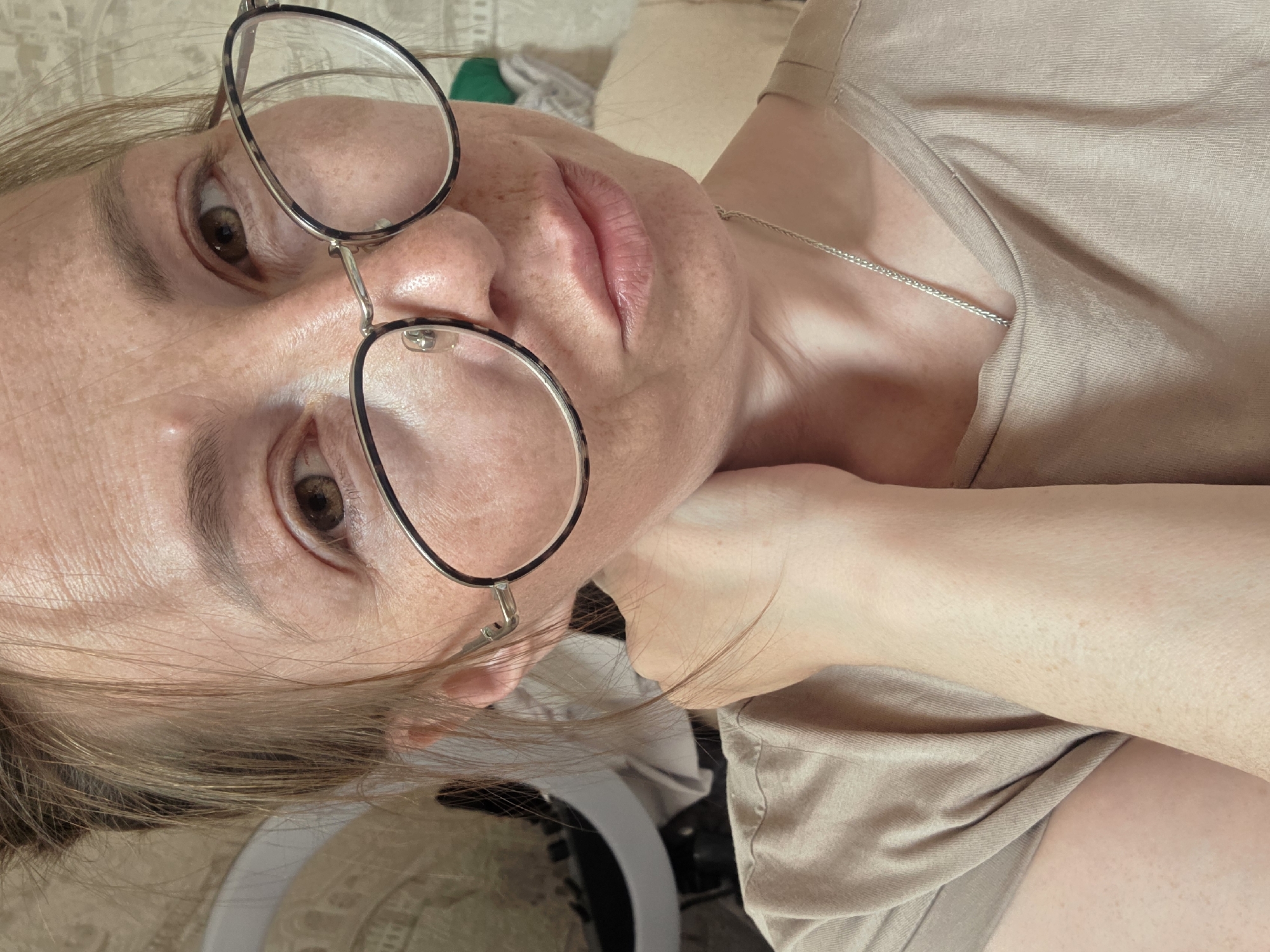 A close-up, rotated image depicts a fair-skinned person wearing glasses and a light brown t-shirt, with a thin silver necklace visible. The person has freckles on their face. The background is blurry. An LED ring light can be partially seen in the lower part of the image. The location is not identifiable.