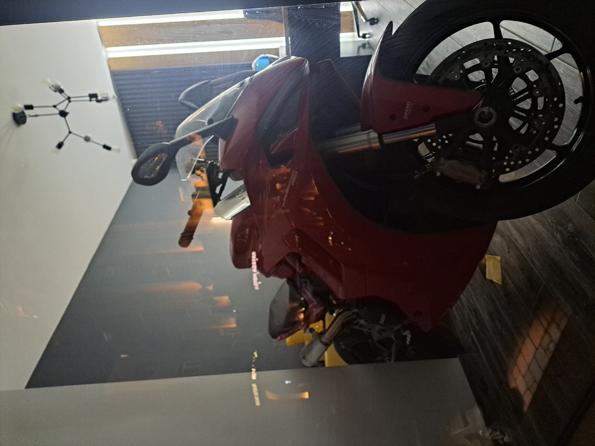The image captures a red Ducati SuperSport 950 motorcycle displayed within a glass enclosure. Reflected in the glass are elements of an interior, including a contemporary light fixture mounted on a white wall. The motorcycle is positioned at an angle, showcasing its rear wheel and brake disc prominently. No people are visible in the image. The location is not discernible from the image alone.
