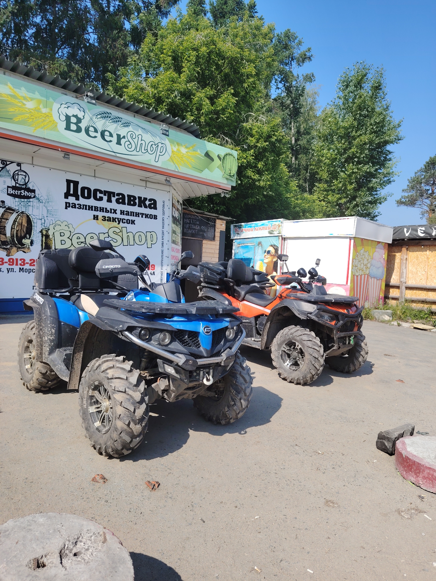 Two all-terrain vehicles (ATVs) are parked on a paved surface in front of a building with multiple signs and a kiosk. The ATV in the foreground is blue and black, with large, knobby tires and a black storage box on the rear. The ATV behind it is orange and black, also with prominent tires and a similar storage box.
The building behind the ATVs has a green awning with the text "Beer Shop" in large, stylized lettering. Below this, a white sign displays "Доставка разливных напитков и закусок" (Delivery of draft beverages and snacks) and another "Beer Shop" logo. To the right of the building, a kiosk with a white roof displays colorful graphics, including ice cream cones. A wooden fence runs along the right side of the scene. Trees with green foliage are visible behind the building and kiosk. The sky is clear and blue. The ground is paved and shows some debris.