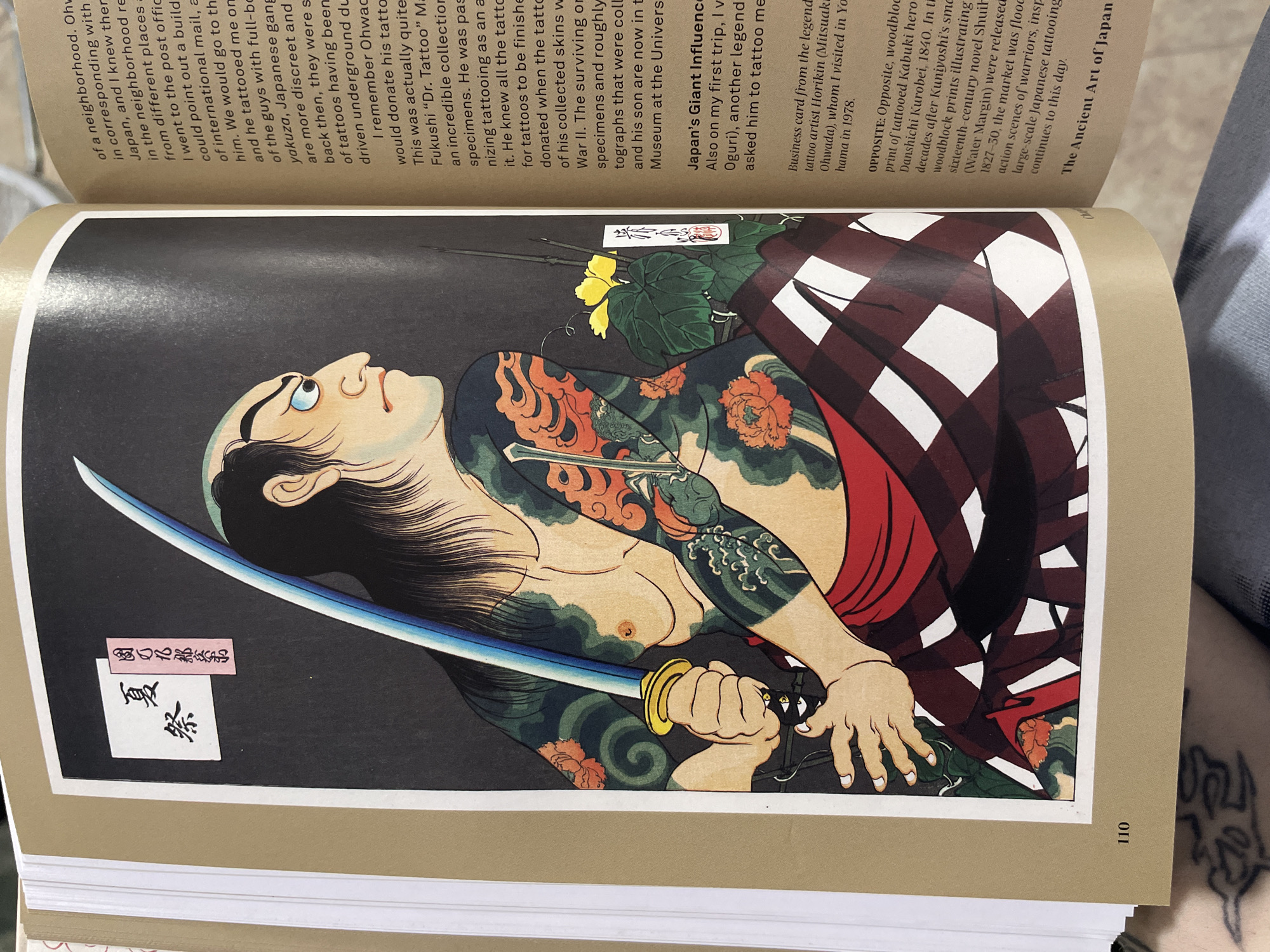 The image shows a page from a book titled "The Ancient Art of Japan," featuring a woodblock print of a tattooed figure, likely a Kabuki hero. The figure is depicted holding a sword and has extensive tattoos on his arms and torso. The book page also includes text discussing the history of tattooing in Japan and its connection to yakuza and the collection of tattooed skins by Dr. Fukushi. The image does not contain information relevant to Buenos Aires, Argentina.