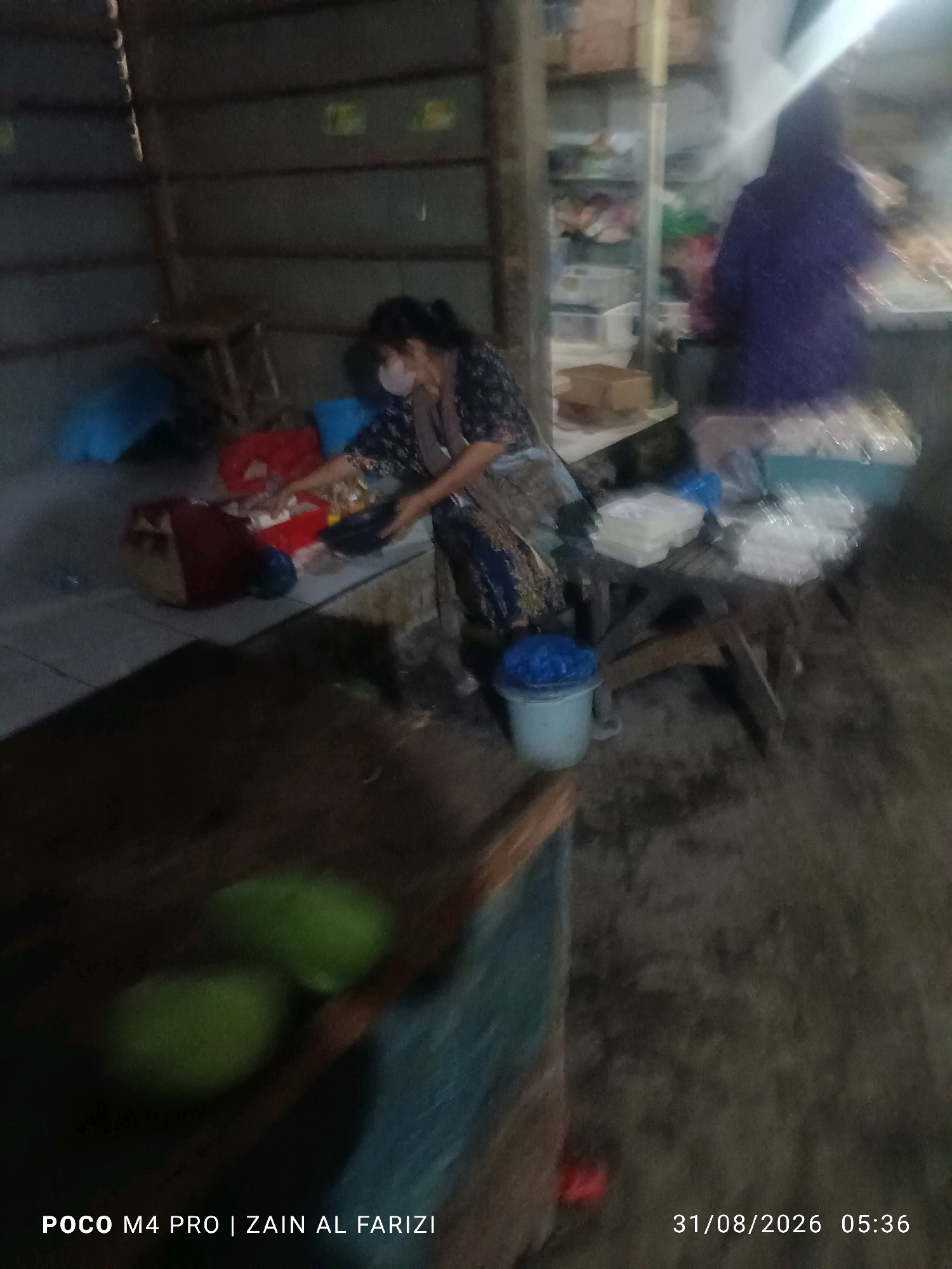 In Arosbaya, Indonesia, a dimly lit indoor market scene unfolds. A woman wearing a mask and a patterned dress is actively engaged, her hands near a red container filled with items. She appears to be either packaging or sorting goods. To her right, another person, dressed in a vibrant purple top, is also partially visible, seemingly engaged in similar activity. The environment suggests a commercial setting with shelves stocked with packaged goods in the background. Several blue and red plastic bags are scattered around the area, along with plastic containers and what appear to be food items. In the foreground, two green fruits, possibly limes or small unripe mangoes, rest on a surface. The timestamp in the lower right corner indicates the image was captured on August 31, 2026, at 5:36 AM, and the watermark at the bottom left identifies the device as a POCO M4 PRO and credits the photographer as ZAIN AL FARIZI. The overall atmosphere is one of a busy, perhaps early morning, market operation.