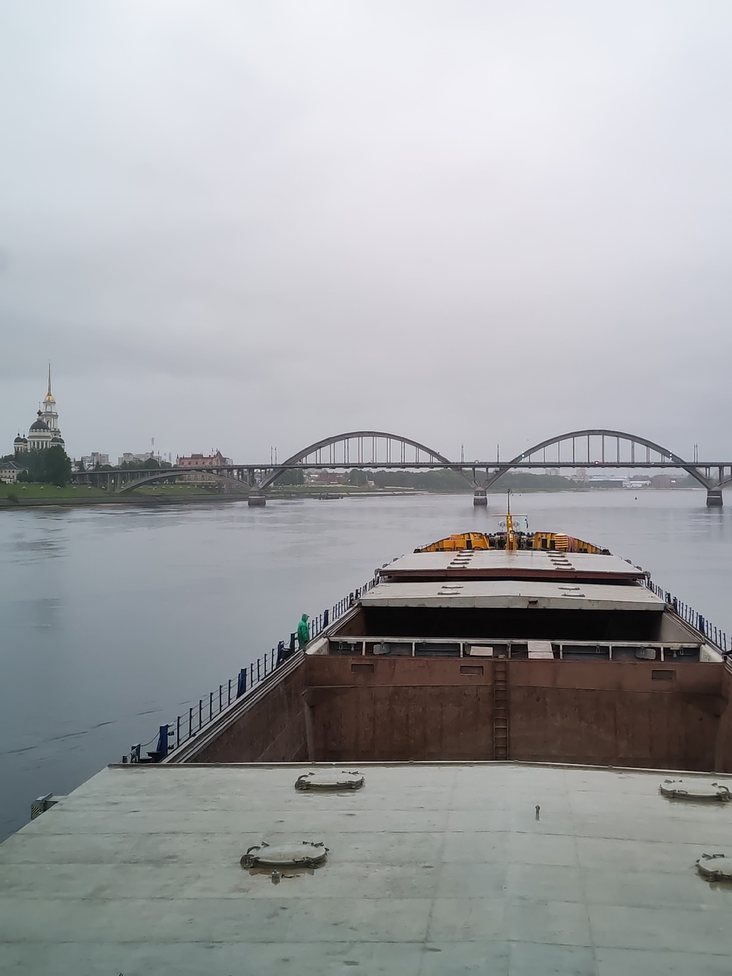 The image depicts a barge moving along a river in Bol'shie Mkhi, Russia, likely the Volga River. The sky is overcast. The barge is primarily gray and brown, with a ladder visible on the interior. A person wearing a green raincoat stands at the edge. In the distance, a bridge with a distinct arched design spans the river, and a church is also visible. The buildings and city infrastructure are situated along the riverbank.