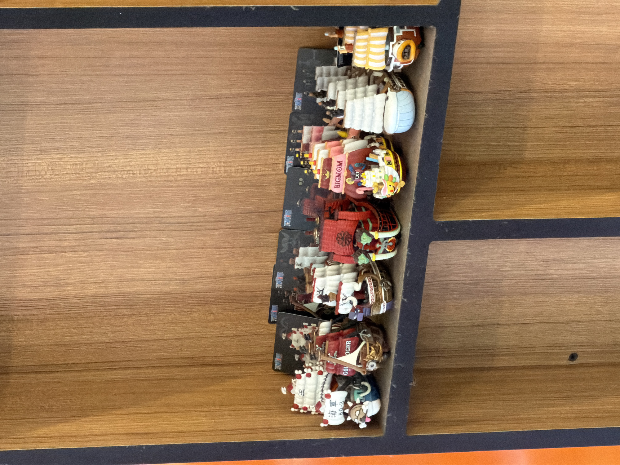 A shelf in a display case holds a collection of small, colorful ship figurines. The shelf is made of wood with vertical grain. Behind the figurines are black cards with printed images, some of which are visible. The figurines themselves are highly detailed, with sails, masts, and decks, and some have characters or faces incorporated into their designs. One figurine is red with a skull and crossbones, another is red with gold accents and what appears to be a flag. A white and blue ship has Japanese writing on its sail. To the right, a pink and yellow ship is labeled "BIGMOM". Further to the right are more white-sailed ships and a yellow and white striped ship. The overall impression is that of a collection of model ships, possibly from a themed franchise, displayed in a retail or private collection setting. The image is taken from a slightly low angle, looking up at the shelf. There is no indication of the specific location within Milan, Italy, as the image only shows the shelf and figurines.