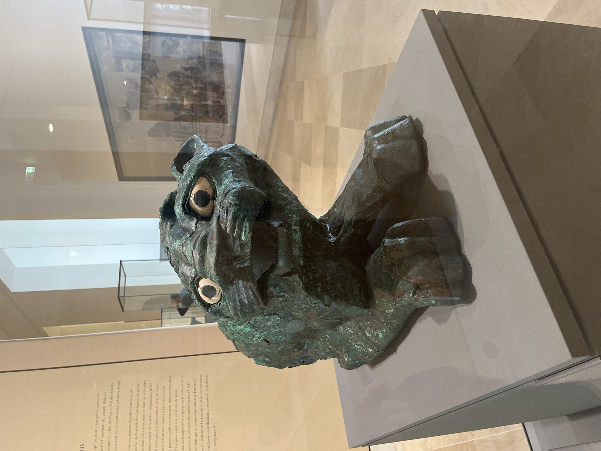 A patinated bronze sculpture, likely a lion or other feline creature, is on display at a museum, possibly in Paris, France. The head and forepaws of the animal are visible. The sculpture is positioned on a grey, stepped platform inside a glass display case. There is a plaque with French text to the left of the sculpture. In the background, a framed artwork hangs on the wall. The museum setting is suggested by the polished stone floors and the reflective surfaces of the glass cases.