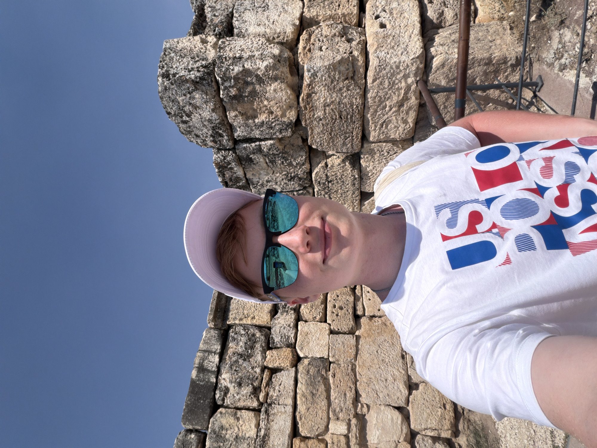 A young Caucasian male is taking a selfie in front of a stone wall under a clear blue sky. He is wearing a white t-shirt with a red, white, and blue graphic design, a white baseball cap, and blue mirrored sunglasses. A beige strap is visible over his left shoulder. The stone wall appears to be ancient, constructed from large, rough-hewn blocks. The setting is outdoors, likely a historical site in Pamukkale, Turkey, given the distinctive stone architecture. A brown railing is visible to the right of the male, suggesting a pathway or viewing area. The overall impression is of a tourist visiting a historical landmark.