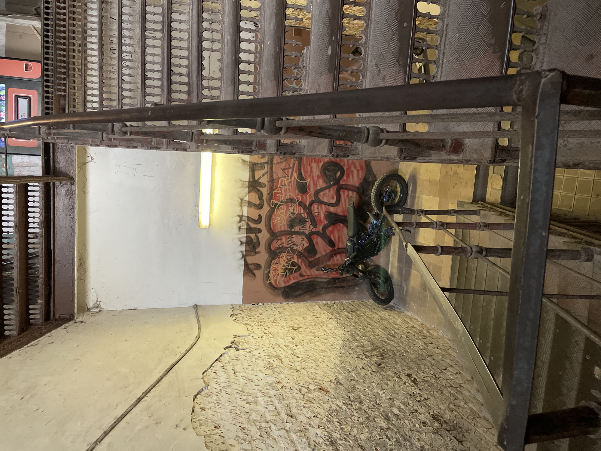 The image depicts an indoor stairwell, possibly in Moscow, Russia. The walls show signs of disrepair, including exposed brick and peeling paint, with graffiti on one wall. A motorcycle is parked in the corner. The stairs are metal with an open grid pattern. The stairwell is illuminated by a fluorescent light fixture. The scene lacks people or visible interaction.