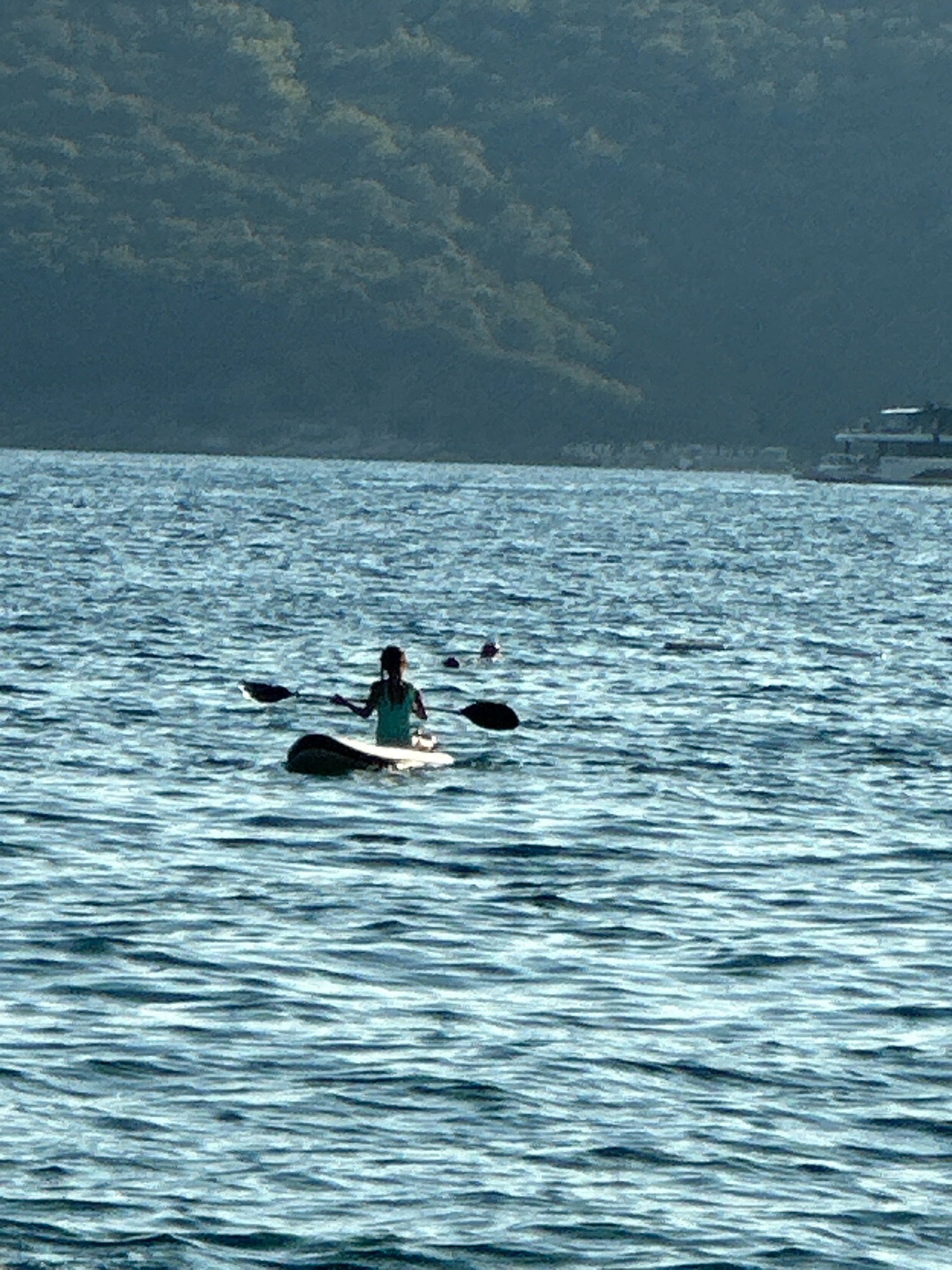 In the city of Boreti, Montenegro, a person is paddleboarding on a body of water. The person is silhouetted against the bright sky and appears to be wearing a turquoise shirt. They are holding a paddle and are positioned on a paddleboard. The water is choppy with visible waves. In the background, a tree-covered hill slopes down towards the water. A white boat is visible in the distance, anchored near the shore. The scene captures a moment of outdoor recreation on a sunny day.