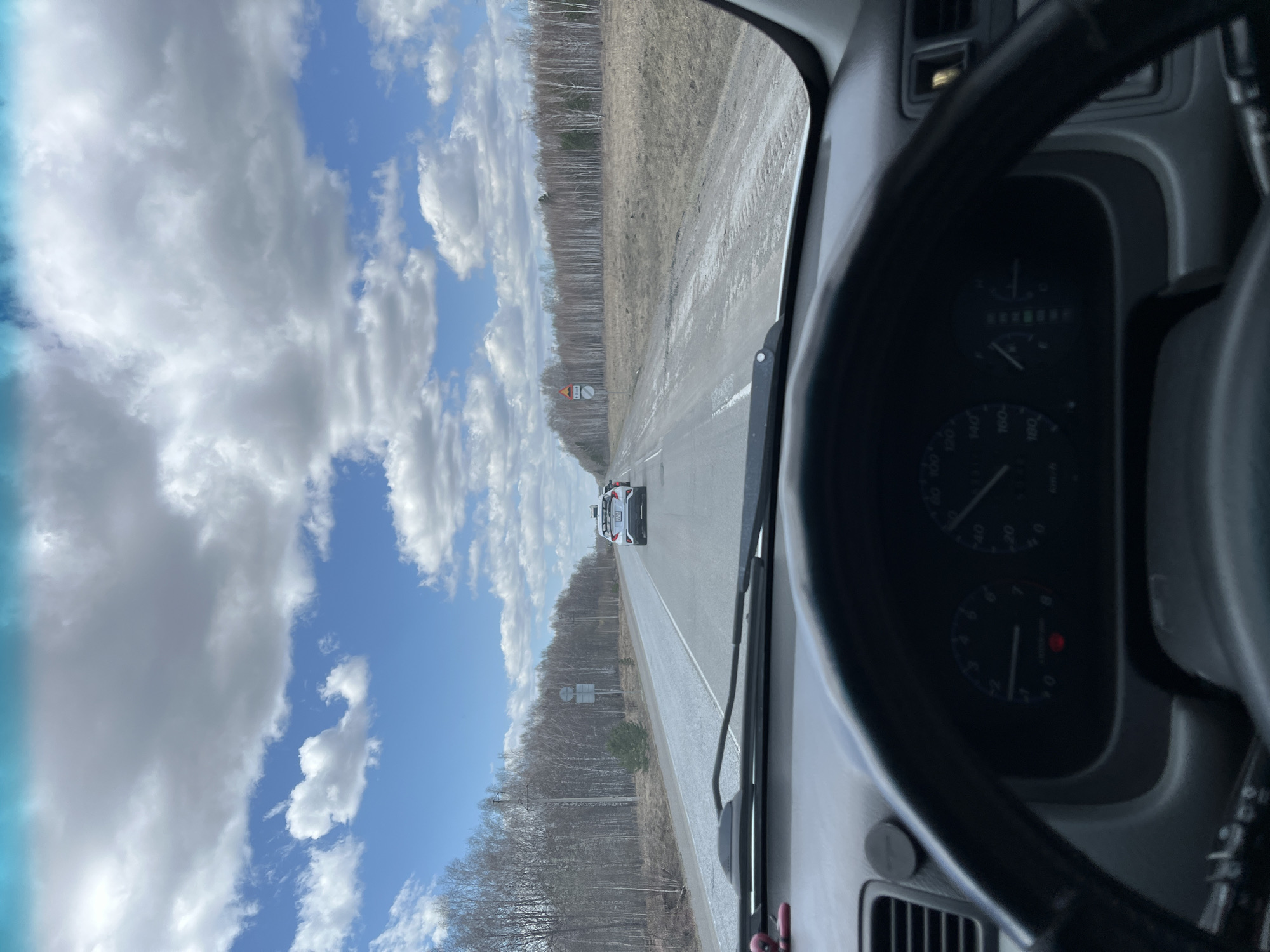 The image shows a view from inside a vehicle, likely a car, traveling on a straight road in Mar'evka, Russia. A white vehicle is traveling ahead. On the left and right sides of the road are sparsely wooded areas. The sky above is blue with scattered white clouds. Part of the steering wheel and dashboard is visible in the foreground.