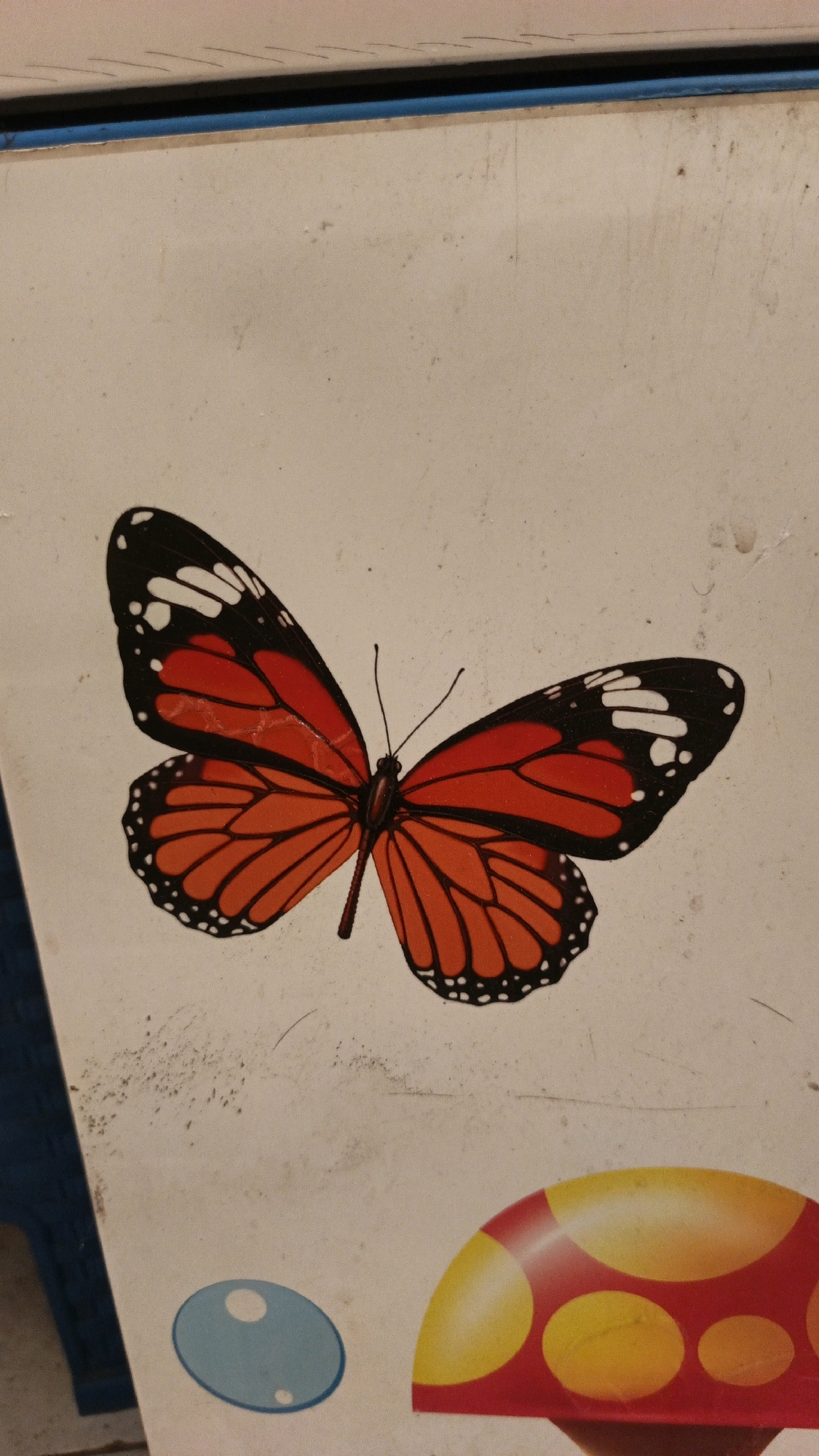 A close-up shot captures a decorative monarch butterfly decal with vibrant orange and black wings, set against a pale, textured surface. The butterfly is positioned centrally in the frame, its wings spread wide, revealing intricate black veining and white spots on the tips. The surface it is on appears to be a textured, off-white material, possibly a painted wall or a piece of furniture. The image also shows a sliver of a blue surface on the left edge and a portion of a red and yellow mushroom graphic at the bottom right. The lighting is slightly dim, casting soft shadows and highlighting the textures of the surface. There are no people or animals present in the image. This image does not provide any information about its location in North Kuta, Indonesia.