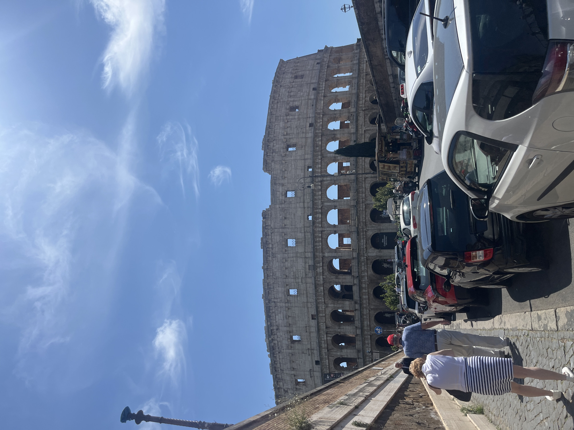 A vibrant outdoor scene captures the iconic Colosseum in Rome, Italy, bathed in bright daylight under a clear blue sky with scattered wispy white clouds. The massive ancient amphitheater, with its characteristic arches and weathered stone, dominates the left two-thirds of the frame.
On the right side of the image, a street is lined with numerous parked cars, including prominent white and black vehicles. Adjacent to the street, a paved walkway or sidewalk occupies the lower right foreground. Several pedestrians are visible on this path, seemingly walking towards the background. Two individuals are most prominent: one person wearing a white top and a navy-and-white striped skirt or dress, and another in a light blue polo shirt, beige pants, and a red hat. Both are seen from behind, suggesting they are moving away from the viewer.
A stone wall or embankment separates the walkway from the street. A dark, ornate streetlamp is partially visible in the bottom left corner. The bright sunlight suggests it is daytime, likely morning or afternoon. Faint text, "Colosseo," is discernible on a sign attached to the Colosseum structure, unequivocally identifying the location. A blue circular road sign with a white arrow is also visible on the street side. The overall scene conveys a typical day at a major tourist attraction in Rome.