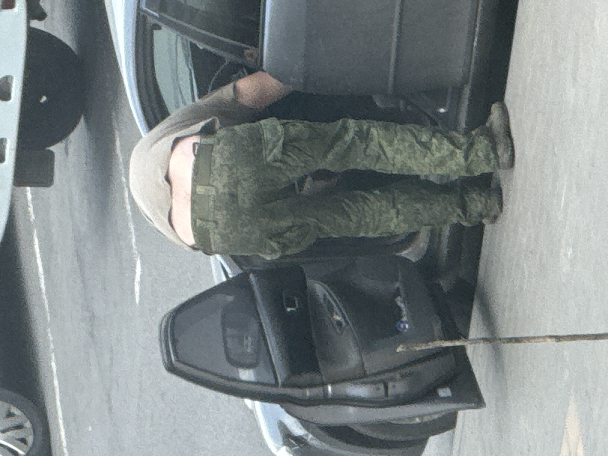A person in camouflage pants and boots is bent over, seemingly exiting a dark-colored sedan with the door open. The setting appears to be an urban area with a paved surface.