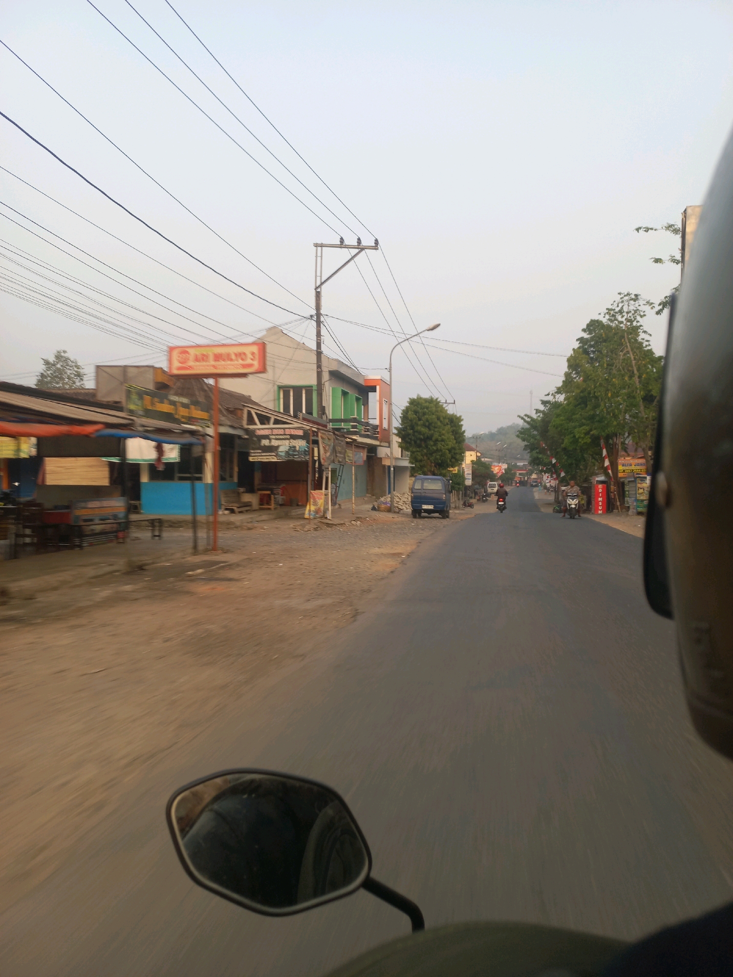 The image shows a street scene in Tirtomoyo, Indonesia, as viewed from a moving motorcycle. In the foreground, the motorcycle's black helmet and a side mirror are visible. The road ahead is paved and appears to be in moderate condition, with some cracks and patches. A blue van is parked on the left side of the road, and other vehicles, including motorcycles, are seen further down the street.
Along the left side of the road are several small businesses or shops. These structures have corrugated metal roofs and appear to be open-air or have open fronts. Signs are visible above some of the shops, with one in orange bearing the text "ARI MULYO 3". There are also tables and chairs set up in front of some of these establishments.
On the right side of the road, more shops are present, and there are trees and utility poles with overhead wires. In the background, a hilly landscape with vegetation can be seen under a hazy sky, suggesting it might be early morning or late afternoon with dust or fog. The overall impression is of a typical street in a rural or semi-urban area in Indonesia.