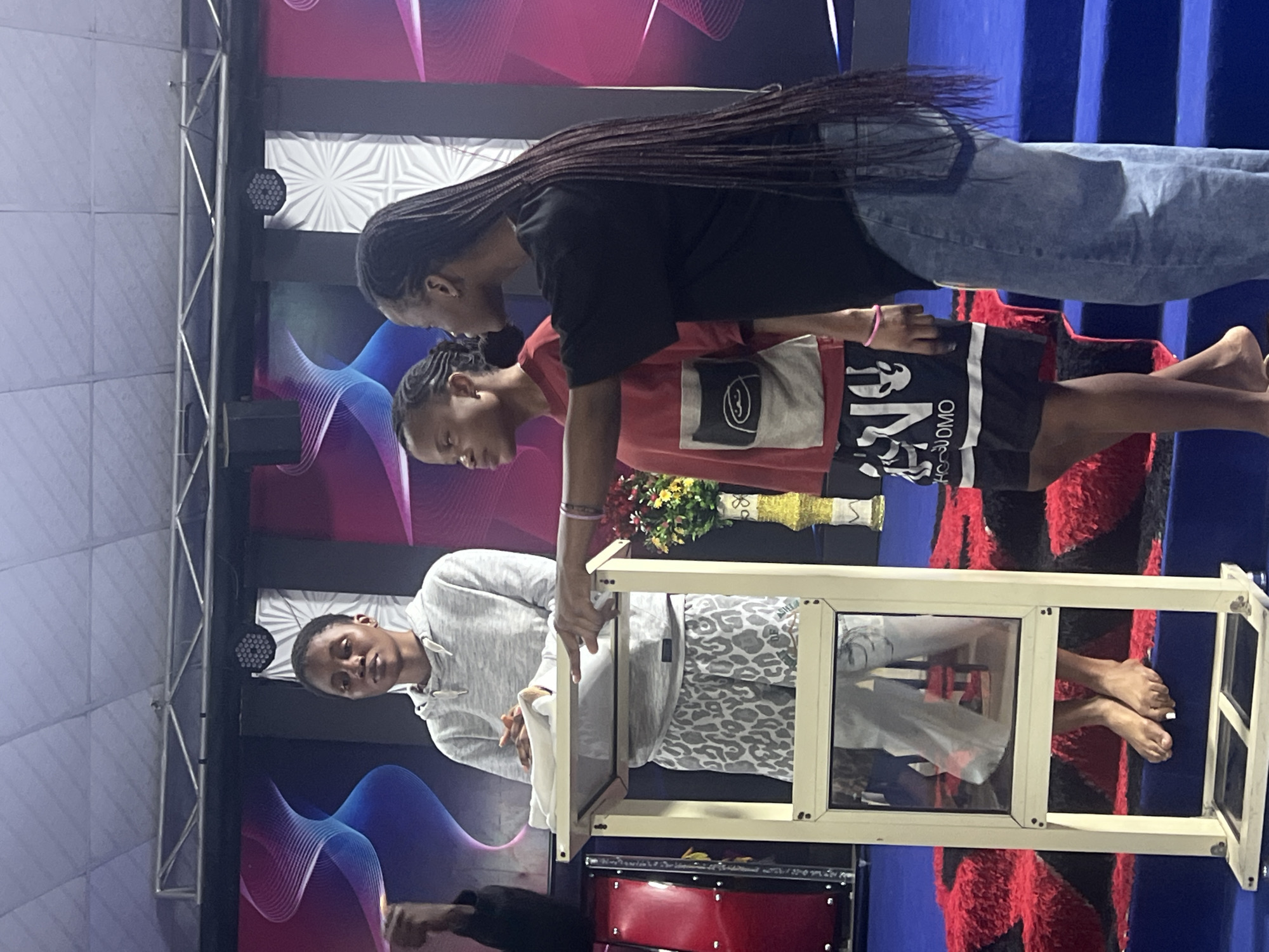 In Nkpor, Nigeria, three young women are standing on a raised platform with a blue carpeted floor and a red rug. The woman on the left is standing behind a transparent podium, holding a white cloth. She is wearing a light grey hoodie and patterned pants. The woman in the middle, facing to her right, is wearing a red t-shirt and black shorts with white lettering. The woman on the right, facing towards the podium, is wearing a black t-shirt and blue jeans. She is pointing towards the podium. Behind them, a stage backdrop features abstract wavy patterns in shades of blue and pink, with lighting fixtures. A red drum is visible to the left of the podium, and a floral arrangement in a yellow vase is to the right. The scene appears to be indoors, possibly a church or event hall.