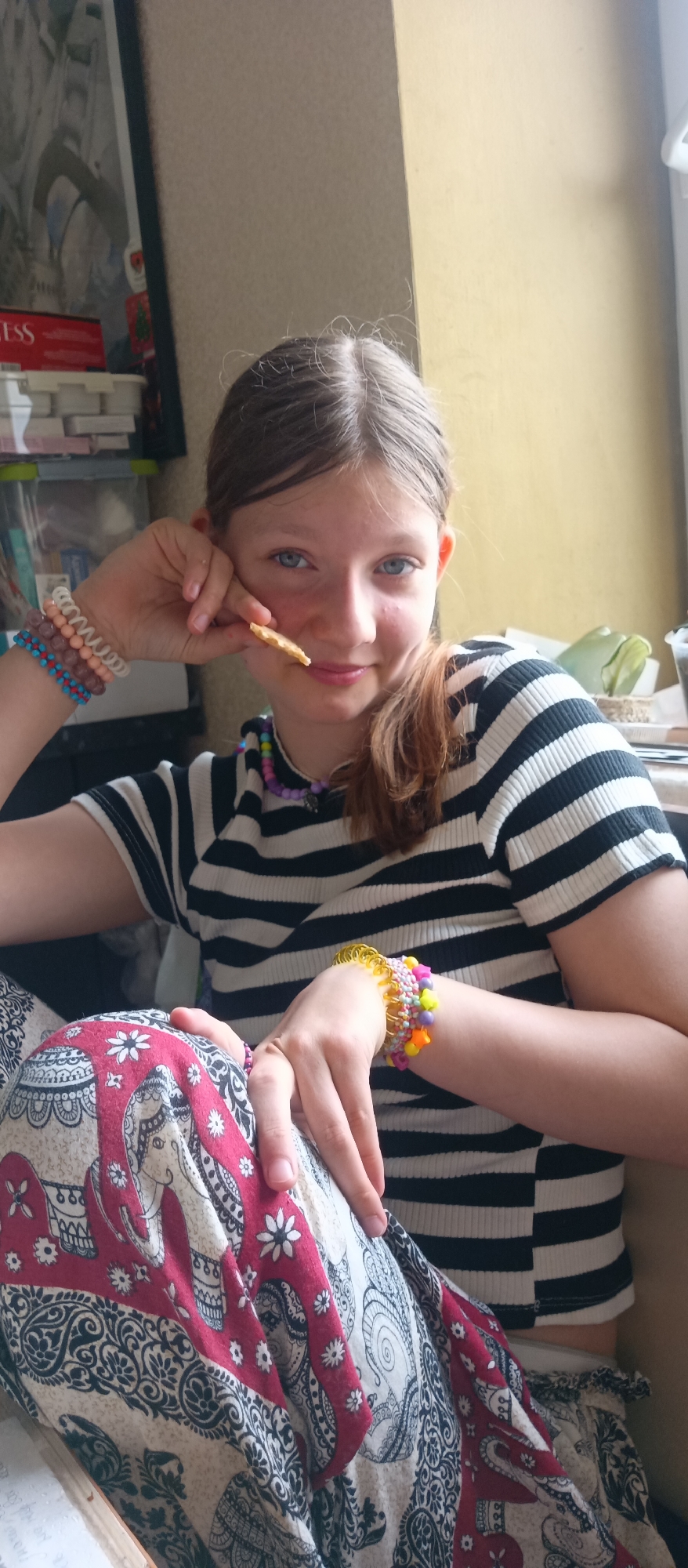 The photograph depicts a young woman, likely in her early to mid-teens, positioned indoors. She is wearing a horizontally striped black and white shirt and patterned trousers featuring an elephant design in shades of red, white, and black. A few bracelets adorn her wrists; the one on her left wrist consisting of blue, brown, and white beads, and on her right wrist consisting of colorful star beads. She is also wearing a necklace made of beads. She is holding a piece of food to her mouth. Her hair is parted and styled in a low ponytail. The background shows a cluttered area with items stacked, a window is visible, along with walls painted in neutral tones. The location is Unknown, Unknown.