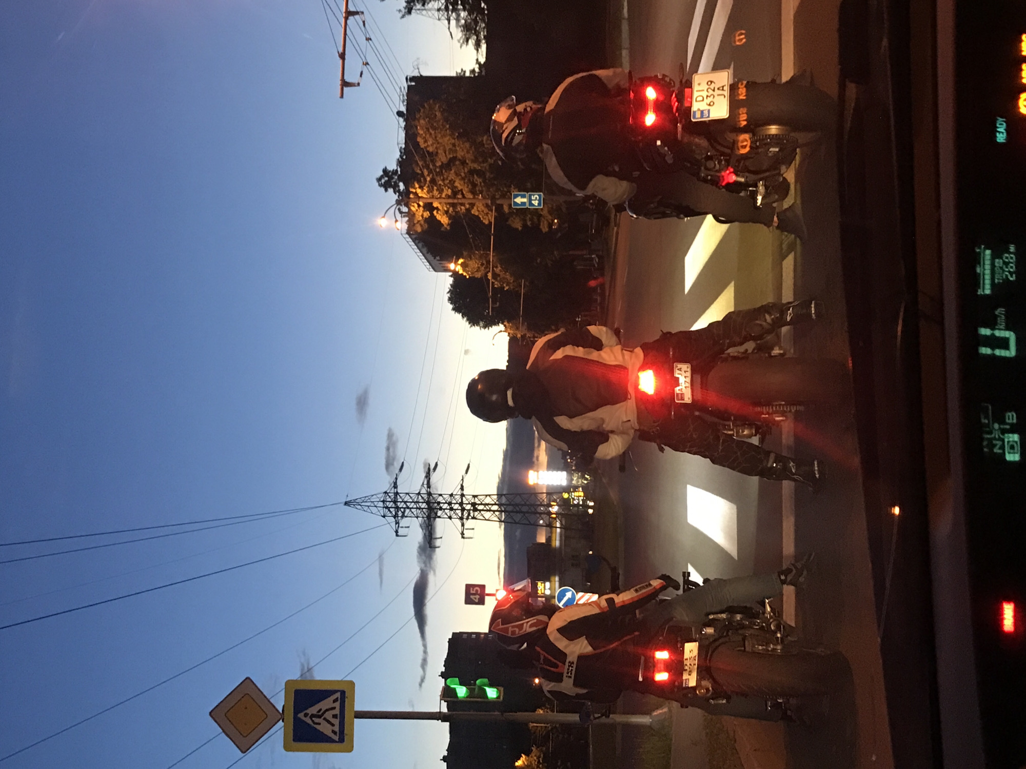 The image captures a scene on a city street at dusk, likely taken from inside a vehicle. The road is lined with streetlights and what appear to be buildings or commercial areas. There are three motorcyclists in the immediate foreground, with their tail lights illuminated. The motorcycles' license plates are partially visible.
Roadside infrastructure is visible, including a signpost displaying road signs. An electrical transmission tower looms in the background. The car's dashboard is also visible, displaying driving information.