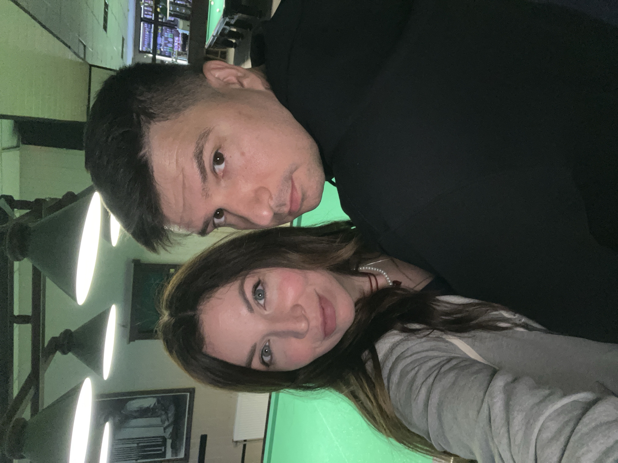 A selfie of two people inside a building, with pool tables visible in the background. A man with short dark hair and wearing a black shirt is positioned behind a woman with long brown hair and wearing a gray shirt. The location appears to be a billiard hall or bar. The city is Unknown, Unknown.