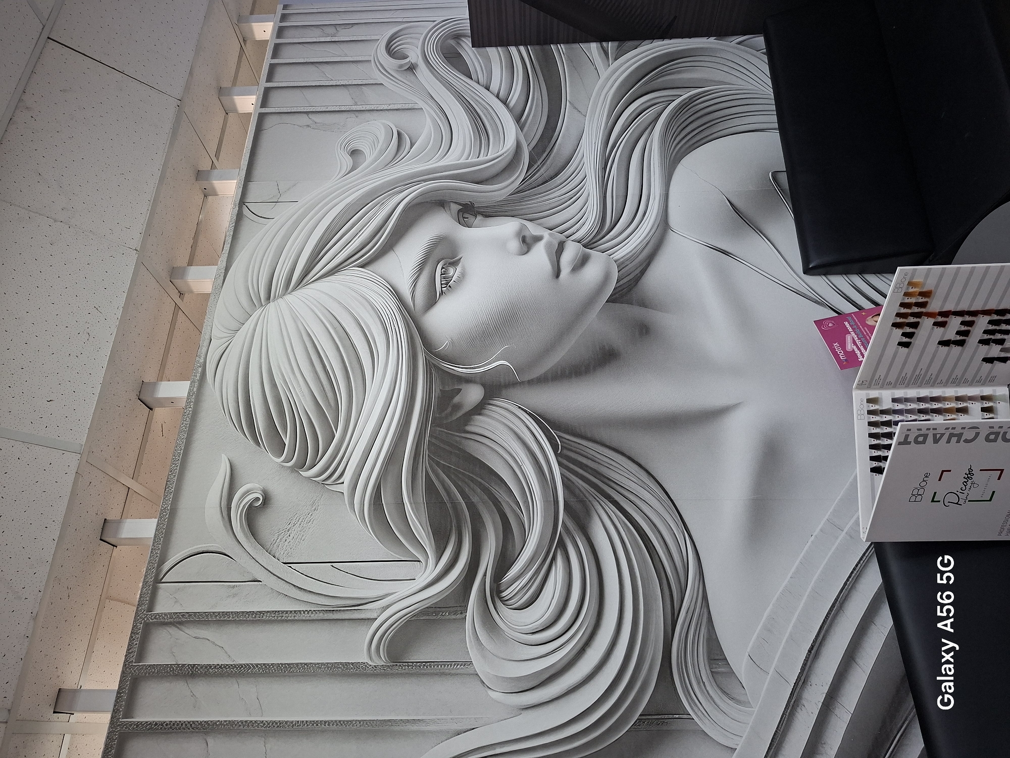 The image shows a monochrome, relief mural of a woman's face and flowing hair against a textured wall. Below the mural, there is a black leather sofa and a display of hair color swatches. A watermark on the lower left corner reads "Galaxy A56 5G". The scene appears to be an interior of a hair salon, possibly located in Chita, Russia, though this location is not explicitly confirmed by visual cues within the image itself.