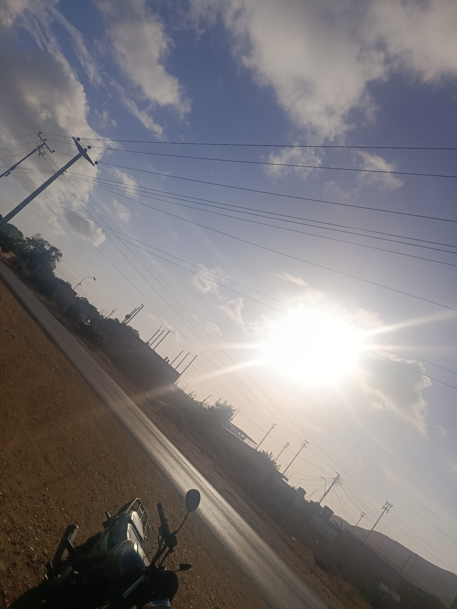 The image depicts a sun-drenched roadside scene in Juan Griego, Venezuela. The view is tilted, showing a road stretching into the distance toward a hazy horizon. A motorcycle is parked on the dirt shoulder in the lower left corner of the frame. Above the road, a series of utility poles carrying power lines crisscrosses the sky. In the middle distance, there are buildings and structures that suggest industrial or commercial activity, including tall vertical cylinders. The sky is a mix of blue and clouds, with the sun a bright focal point.
