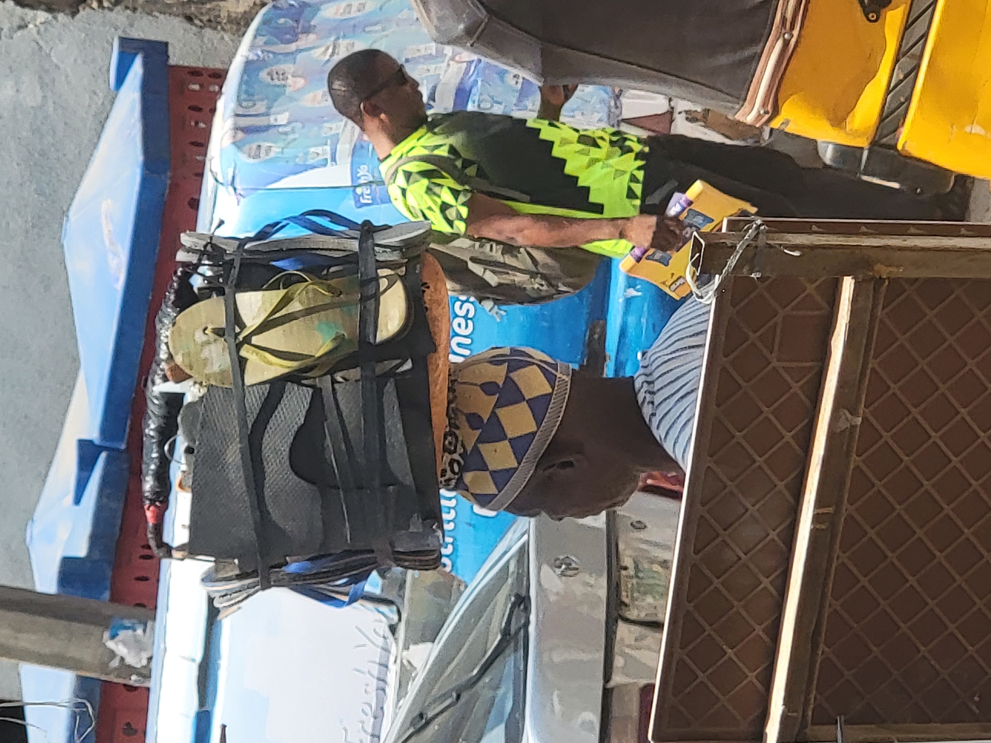 In the city of Maiburiji, Nigeria, two men are seen standing beside a commercial vehicle. One man, wearing a shirt with a geometric green and black pattern, holds a small, yellow rectangular object and appears to be interacting with someone on the vehicle. The other man is wearing a hat with a blue and gold diamond pattern. The vehicle, which appears to be heavily loaded with various items, including packaged goods, has a stack of sandals secured on top. The environment seems to be an urban setting with parts of buildings and other vehicles visible in the background.