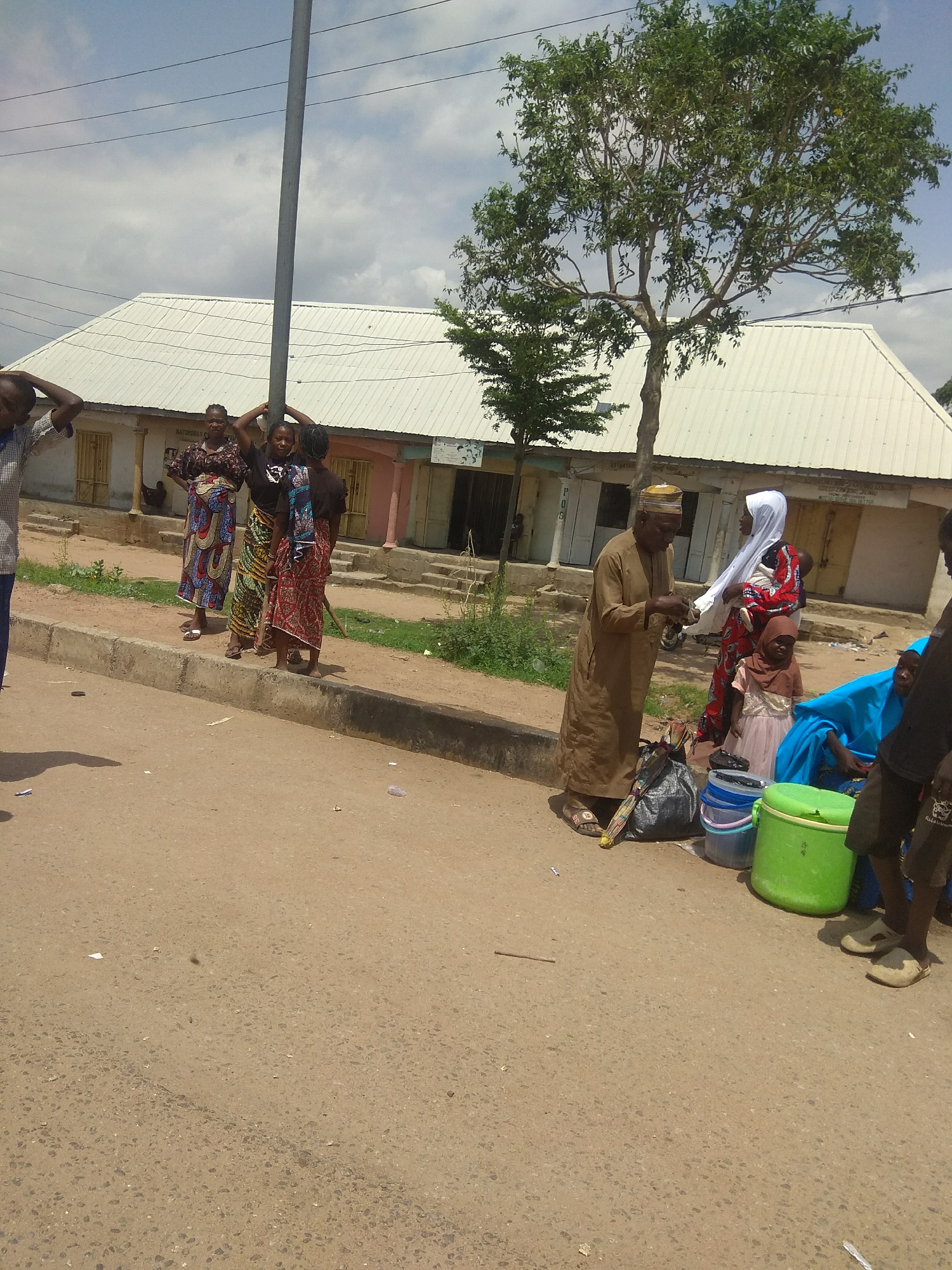 The media file depicts an outdoor scene in Jalingo, Nigeria, under a cloudy sky suggesting daytime with possibly overcast weather. A group of people, including men, women, and children, are gathered near a building with a corrugated metal roof. The building appears to be a shop or a series of shops, with visible doors and windows.
In the foreground, several individuals are interacting. An older man in a tan robe and cap is engaged in conversation with a woman wearing a white hijab and a colorful patterned dress, who is holding a baby. Nearby, a young girl in a pink dress and brown headscarf stands. A large green cooler, blue and white buckets, and a black bag with an umbrella are placed on the ground, possibly indicating a market setting or a stop for goods.
Further down the street, a group of three women are standing together, some with their hands on their heads, their clothing suggesting traditional Nigerian attire. Another person, partially visible on the left, appears to be a young person. The ground is a dirt road with some scattered debris. A utility pole stands prominently in the background.
There is some visible text on the building, including what appears to be "NATURALS CH." The overall atmosphere seems to be one of daily life and social interaction in a community. The gathering of people and the presence of goods suggest a potential market or a public gathering area.