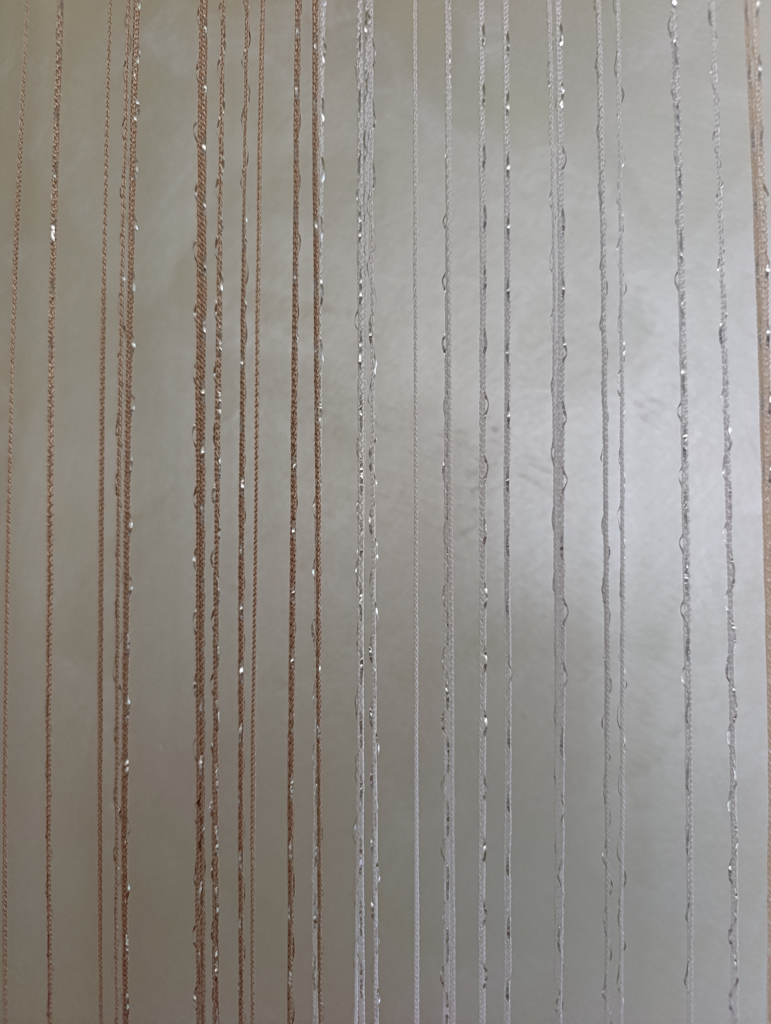 The image shows a close-up view of a beaded curtain or string decoration against a textured, off-white wall. The curtain is composed of multiple vertical strands of thin, braided threads. Some strands are a light tan or copper color, while others are white or silver. Interspersed along the strands are small, reflective elements that appear to be clear or silver beads or sequins, catching the light and creating small glints. The strands hang parallel to each other, creating a uniform, vertical pattern. The background wall has a subtle, mottled texture, suggesting a painted or wallpapered surface. No people or specific locations are visible.