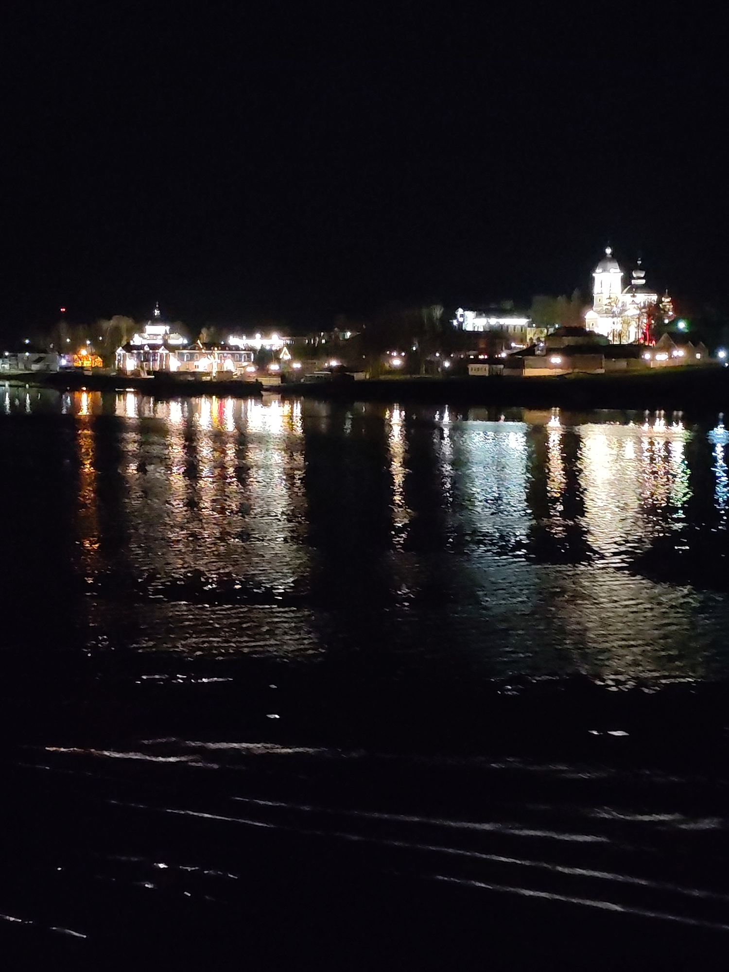 The image depicts a nighttime scene in Nesterovo (Slobodskoe S/P), Russia, with a body of water (likely a river or lake) in the foreground. Illuminated buildings are visible on the opposite bank, with their lights reflecting on the water's surface. One notable structure on the right side of the frame features a distinctive dome, indicating a possible religious building or church. The lighting is dim, with the artificial lights from the buildings creating the primary source of illumination. The water appears dark with subtle ripples and reflections.