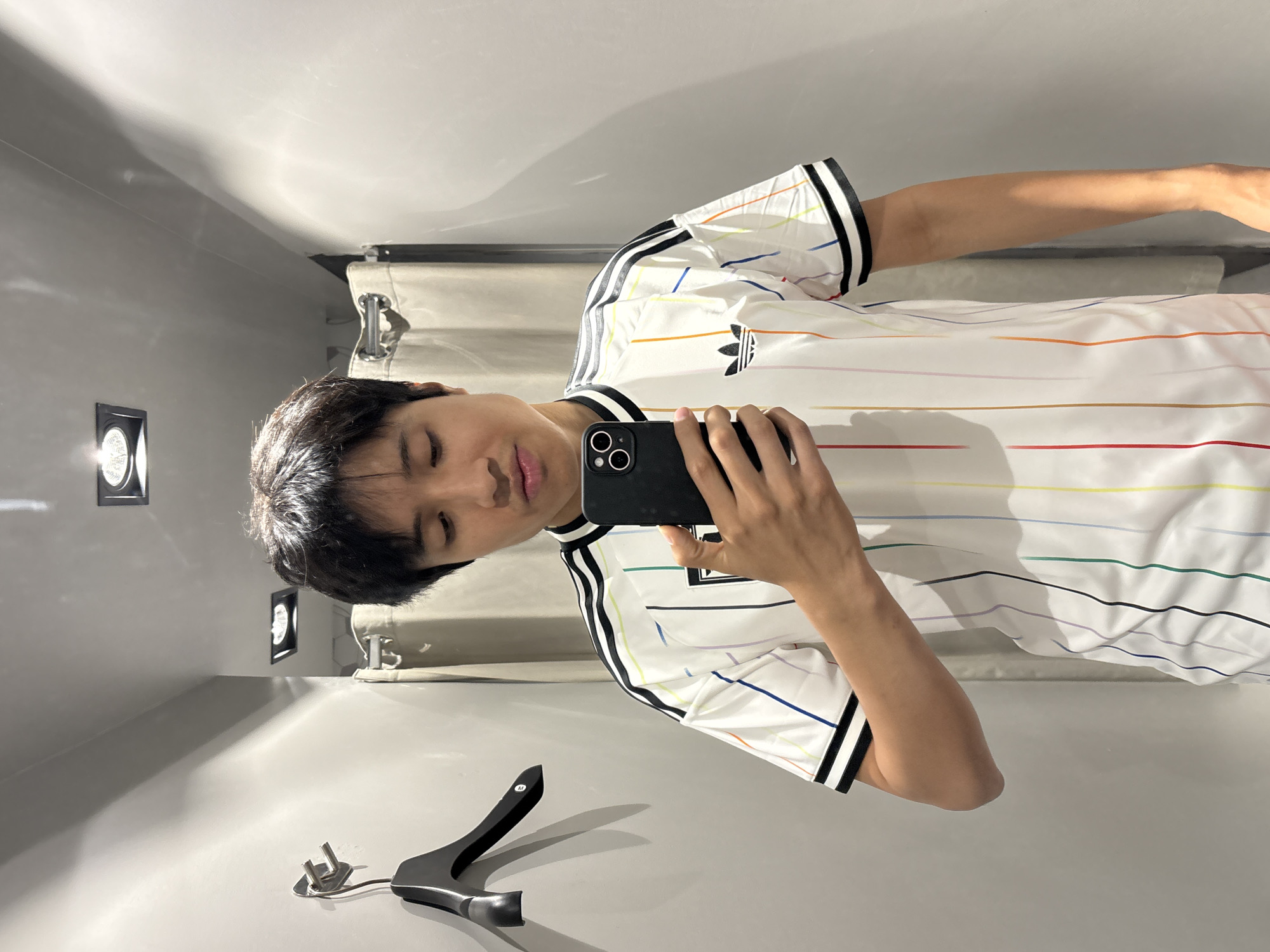 A person stands in a changing room, taking a selfie with a smartphone. The individual wears a white Adidas shirt with black trim on the sleeves and collar, along with horizontal stripes of various colors across the fabric. The changing room has gray walls and recessed lighting fixtures. Clothing racks with gray curtains are visible in the background. A black hanger with metallic components rests on a ledge. The location is plausibly a retail store in a city, but specific details to identify São Paulo, Brazil, are not apparent in the image.