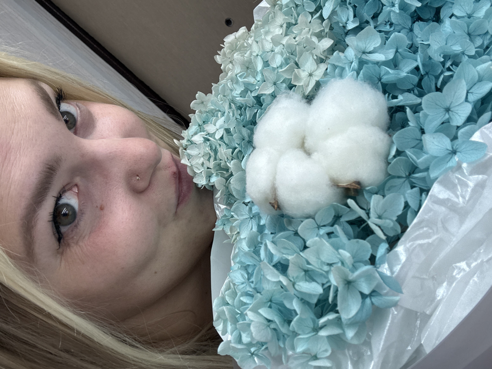 The image is a close-up, vertically oriented photograph of a young woman's face and a bouquet of dried blue hydrangeas and cotton. The woman, with blonde hair and fair skin, is looking towards the camera with a neutral expression. Her right eye is visible, showing a green iris and dark eyelashes. She has a small nose piercing. The bouquet of flowers is on the right side of the image, with soft blue petals and fluffy white cotton balls. The background is out of focus, but appears to be an indoor setting with wooden elements. The lighting suggests it could be daytime. No text is visible in the image. The overall mood is serene and delicate.