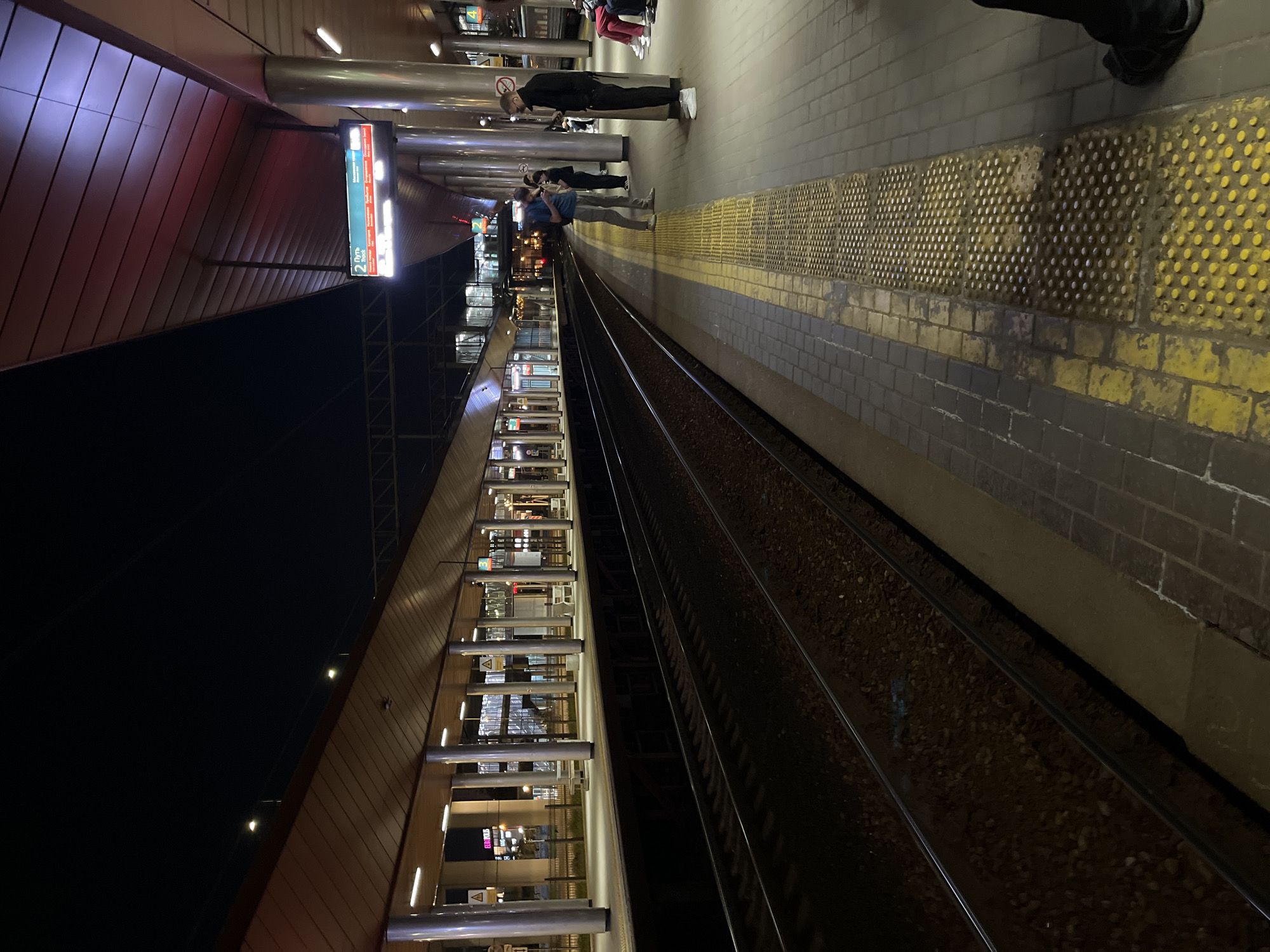 A night time view of a train station platform in Moscow, Russia. The platform is illuminated by overhead lights and a digital sign above the tracks indicates "2 Путь" (Track 2) with train destinations. Several people are standing on the platform, some looking at their phones, while others wait. The train tracks run along the left side of the image, with ballast beneath them. The platform itself is paved with tiles and features tactile paving for the visually impaired. In the background, the station's architecture is visible, including supporting columns and a covered roof.