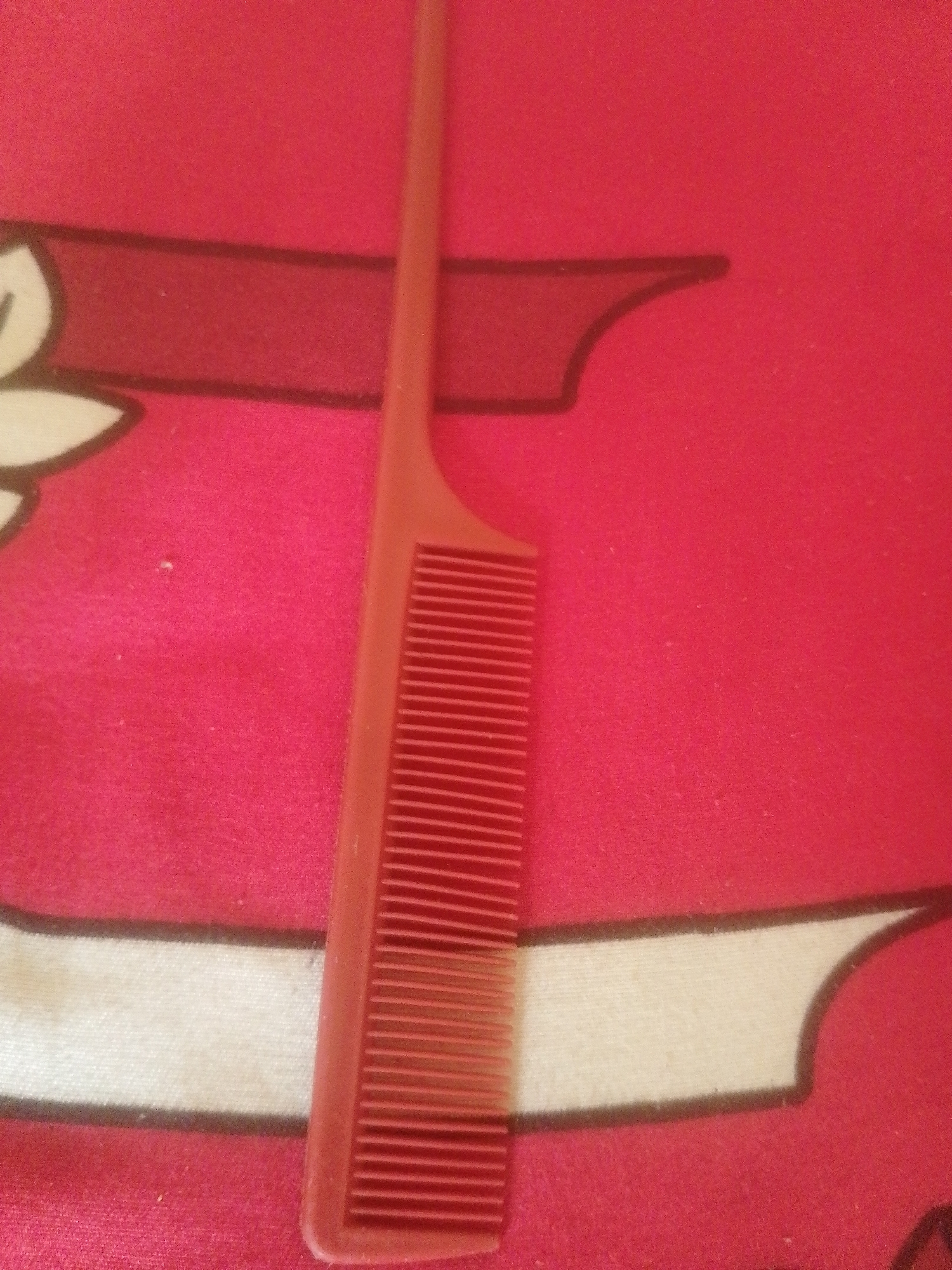 The image displays a close-up, top-down view of a red comb resting on a red patterned fabric surface. The comb is positioned vertically, with its teeth facing downwards. The fabric has a textured appearance and features a repeating pattern of dark red outlines and white shapes. The lighting appears to be artificial, casting subtle shadows. There is no indication of the location being Majemu, Nigeria, or any specific urban environment. The scene solely consists of the comb and the fabric.