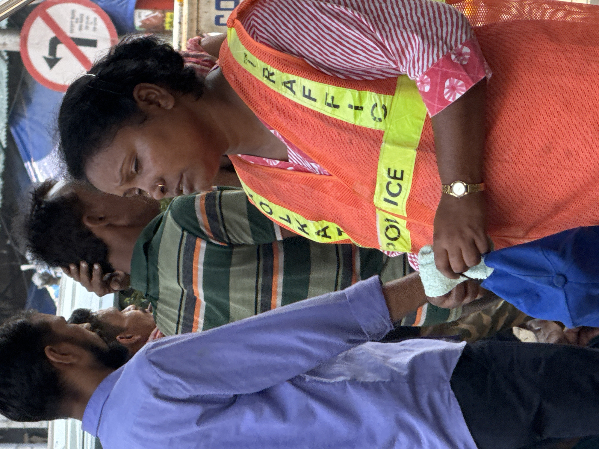 The image shows a Kolkata, India, traffic police officer in a red-and-white striped shirt, a red mesh vest with yellow reflective strips labeled "TRAFFIC POLICE," and a gold watch on her left wrist. She is interacting with a man in a light purple shirt and black pants, holding a light-colored cloth and a blue hat. Behind the officer and the man is another man in a green-and-brown striped shirt. A traffic sign prohibiting left turns is visible in the upper left corner of the image. The environment appears to be a busy street scene.