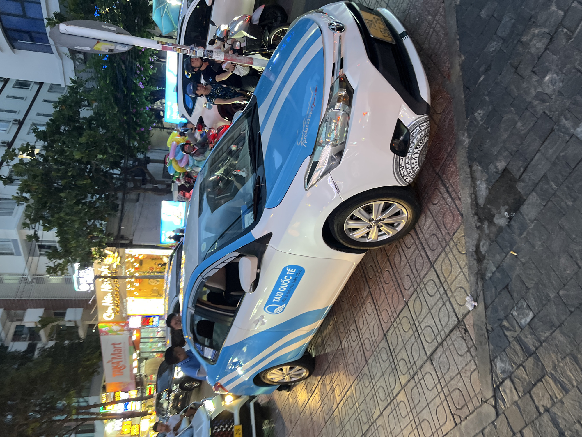 A white and blue taxi with the logo "TAXI QUỐC TẾ" is parked on a tiled sidewalk in Nha Trang, Vietnam. The taxi has a blue stripe design on its hood and sides. In the background, there are shops with bright lights, trees, and other vehicles, including motorbikes. Several people are visible, some riding motorbikes and others standing near the taxi. The scene appears to be a busy street in a commercial area of the city.