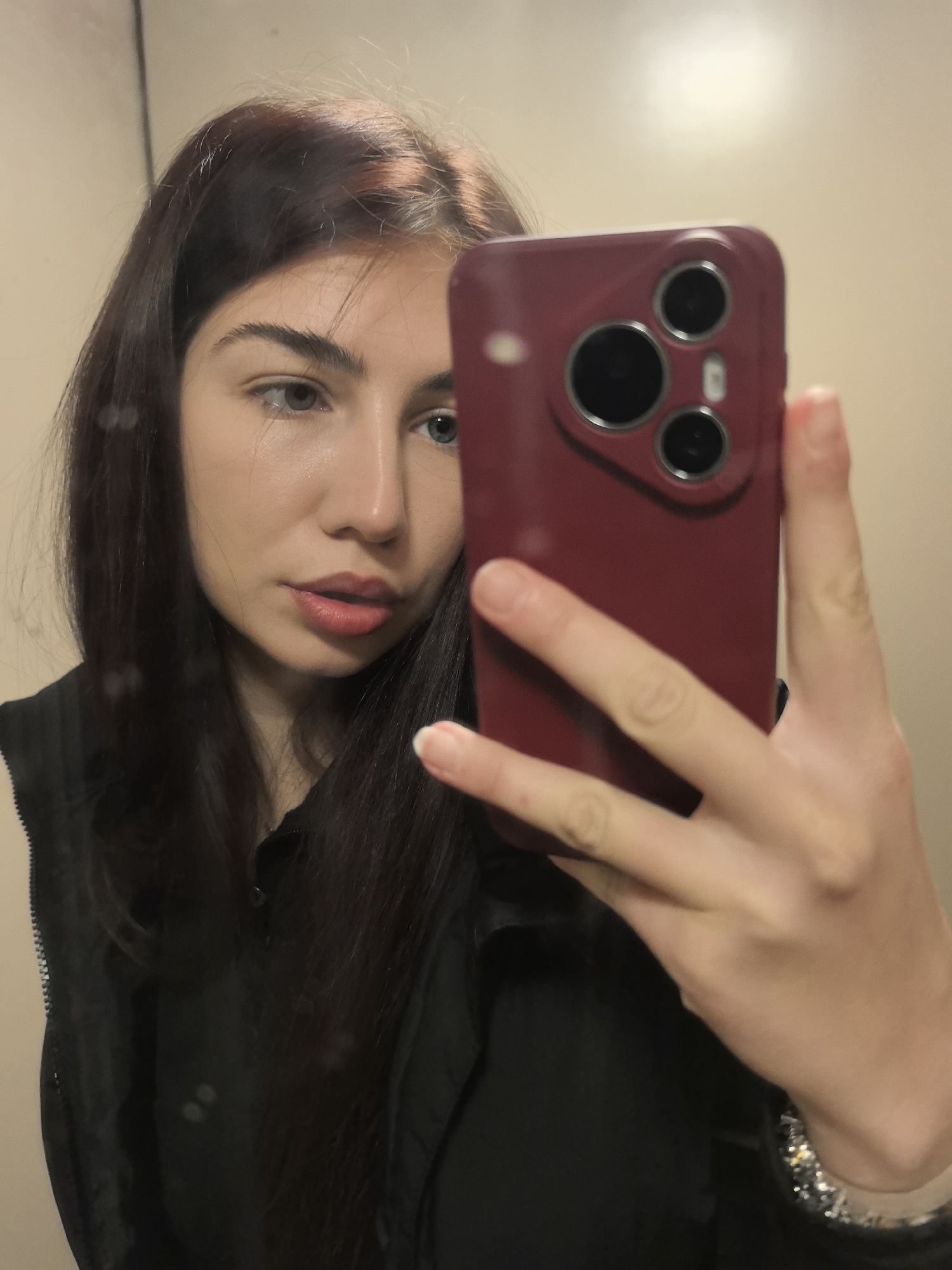 A young woman is taking a selfie in what appears to be an indoor setting with beige walls. She is holding a burgundy-colored smartphone with three camera lenses in front of her face, partially obscuring it. The woman has dark hair with hints of auburn and is wearing a black top or vest. She has fair skin, light-colored eyes, and slightly parted lips. A silver bracelet is visible on her wrist. The location is not discernible from the image.