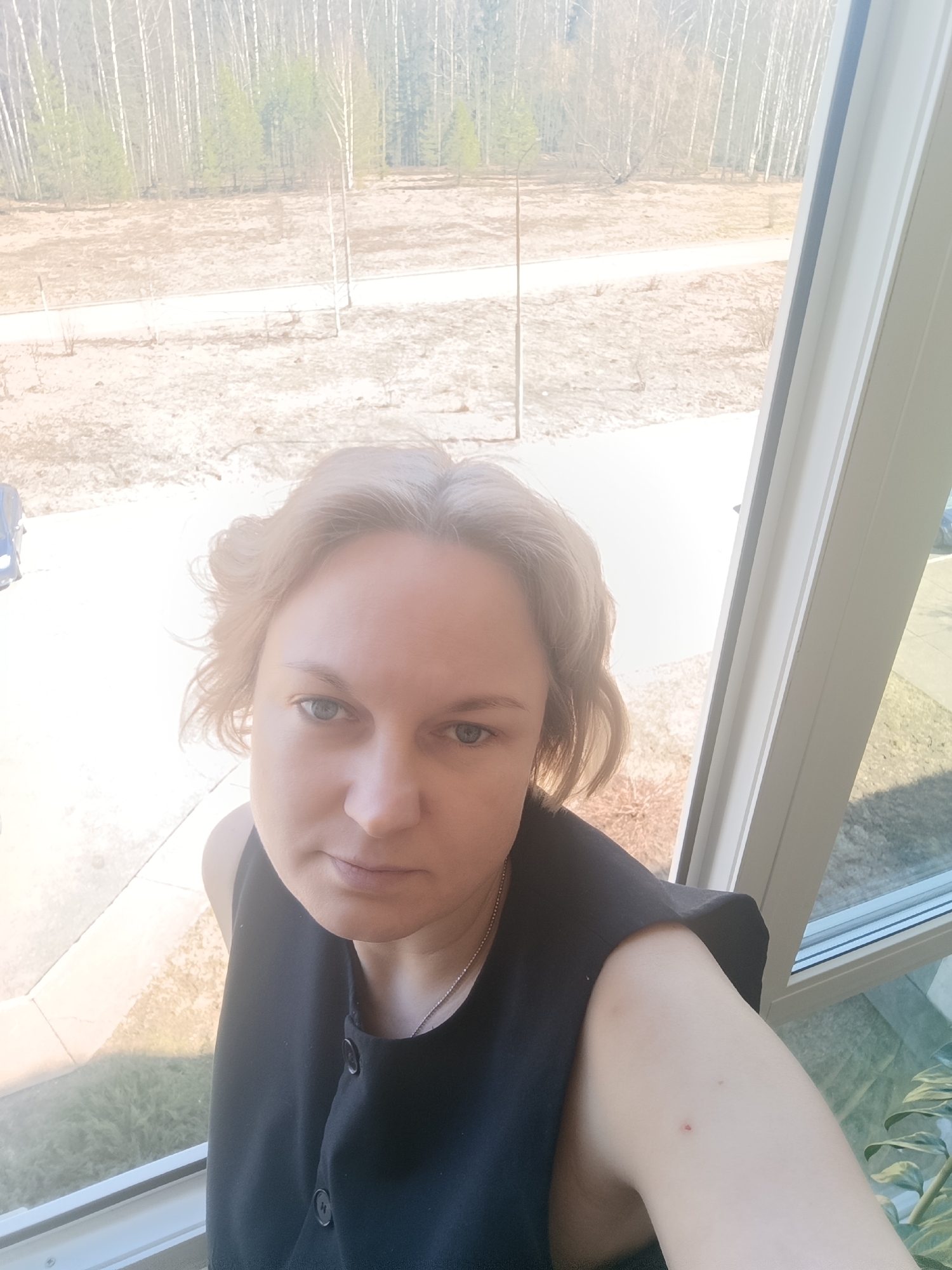 A woman with short, light blonde hair is posing for a selfie near a window. She has fair skin and blue eyes, and is wearing a sleeveless black top with buttons. A silver chain necklace is visible around her neck. Outside the window, there is a view of bare trees, a light-colored ground surface, and a walking path. Based on the surrounding environment of barren trees and pale colored ground, the picture appears to be taken in the city of Марковский, Russia, which experiences a tundra climate.
