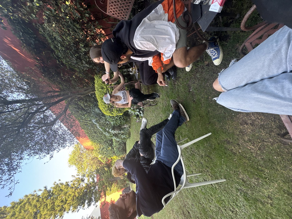 Several individuals are gathered outdoors. One wears a white Adidas tank top and a baseball cap. Another person smokes a cigarette, seated at a table with a pack of Marlboro cigarettes. Other individuals are seated around the table, with one wearing light wash denim jeans and another wearing brown shoes. There is a mix of green foliage and trees in the background.