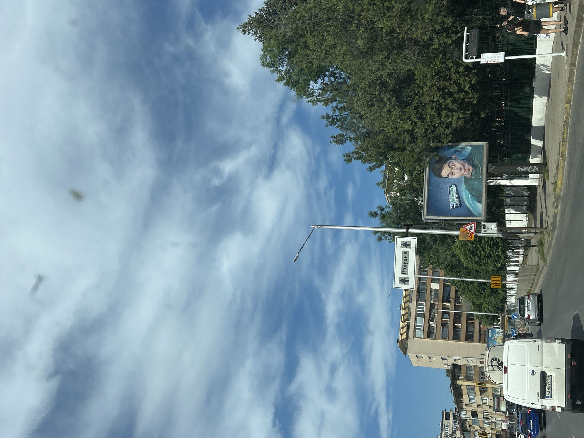 Captured from a vehicle on a street in Sofia, Bulgaria, the image features a bright, partly cloudy sky dominating the upper portion.
A roadside billboard displays an advertisement with a human face and a product, placed near leafy trees. Traffic and buildings align the right side of the image, including white vans and vehicles with visible license plates. Street signs are also present, indicating place names in Cyrillic. In the top-right corner, several people are visible on a walkway near the street.