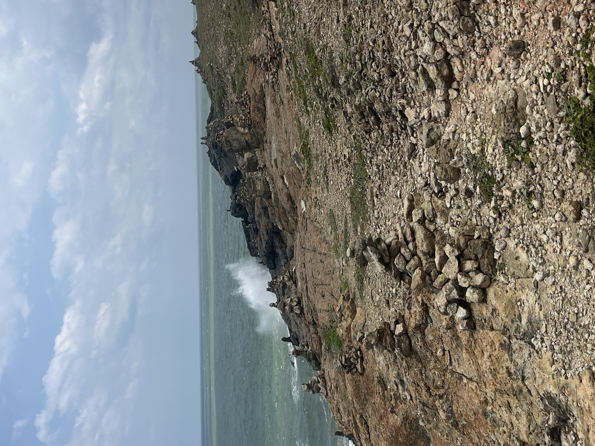 The image depicts a rocky coastline with the ocean in the background. Numerous cairns, or stacked stones, are visible on the rocks and the grassy slopes leading to the sea. The sea is a muted green, with a wave crashing against the rocks on the left side of the frame, creating white foam. The sky is overcast with a mix of white and grey clouds. The terrain is uneven and covered in loose stones and sparse vegetation. The scene is likely a natural landmark or tourist attraction in Mui Ne, Vietnam, possibly related to spiritual practices or a form of art. There are no people visible in the image.