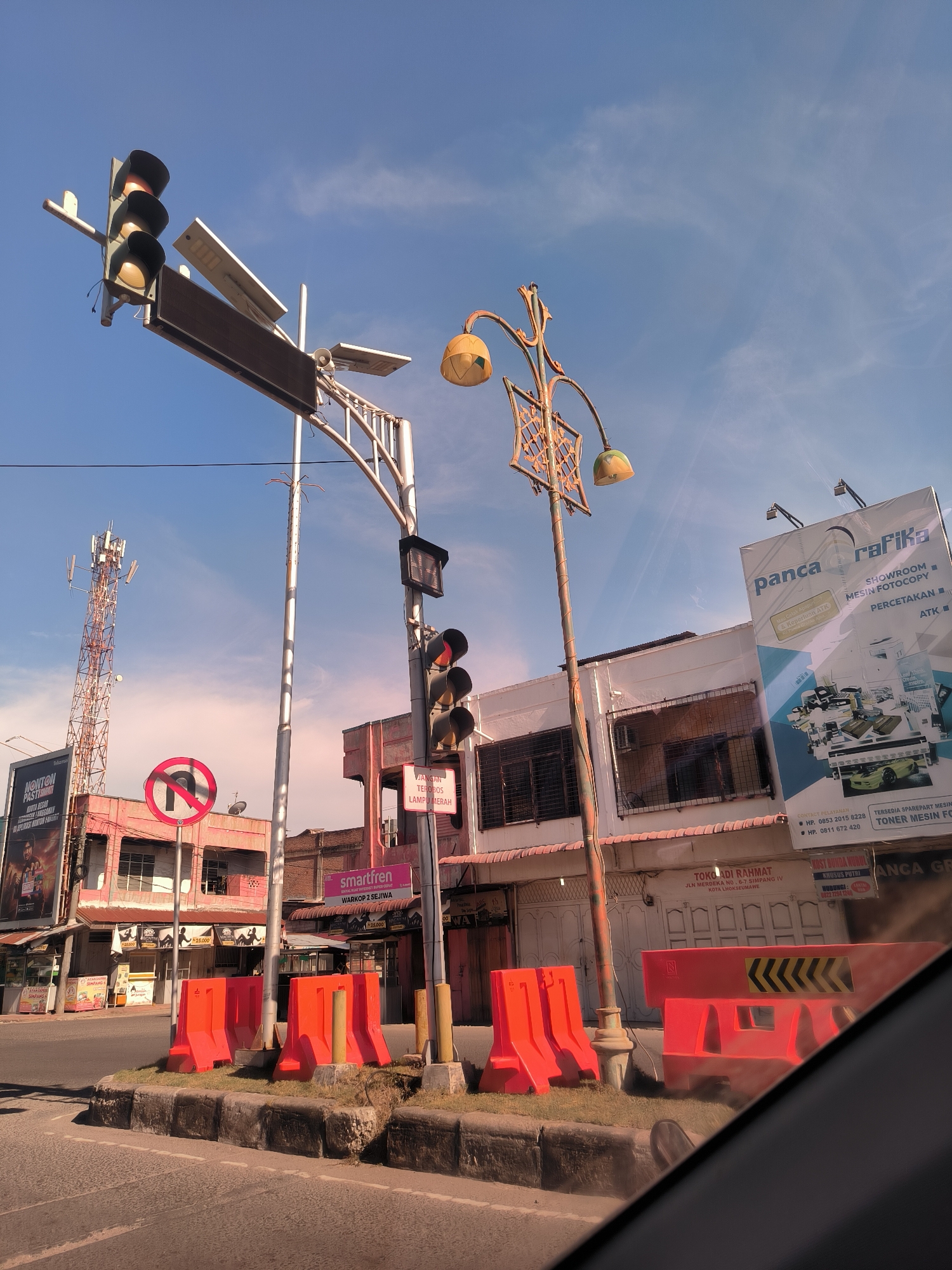 The image captures a street scene in Lhokseumawe, Indonesia, near the intersection of Jalan Merdeka and Simpang IV. The sky is blue with light clouds. Traffic lights stand on poles on a median strip, accompanied by orange traffic barriers. A decorative street lamp is also visible. A cell tower rises in the background behind a pink building. Several commercial billboards are present, advertising various businesses, including Panca Rafika (office equipment), Smartfren (telecommunications), and local restaurants. The image is taken from inside a vehicle, indicated by the interior dashboard or frame visible at the bottom. The overall impression is of a busy urban intersection during daylight hours.