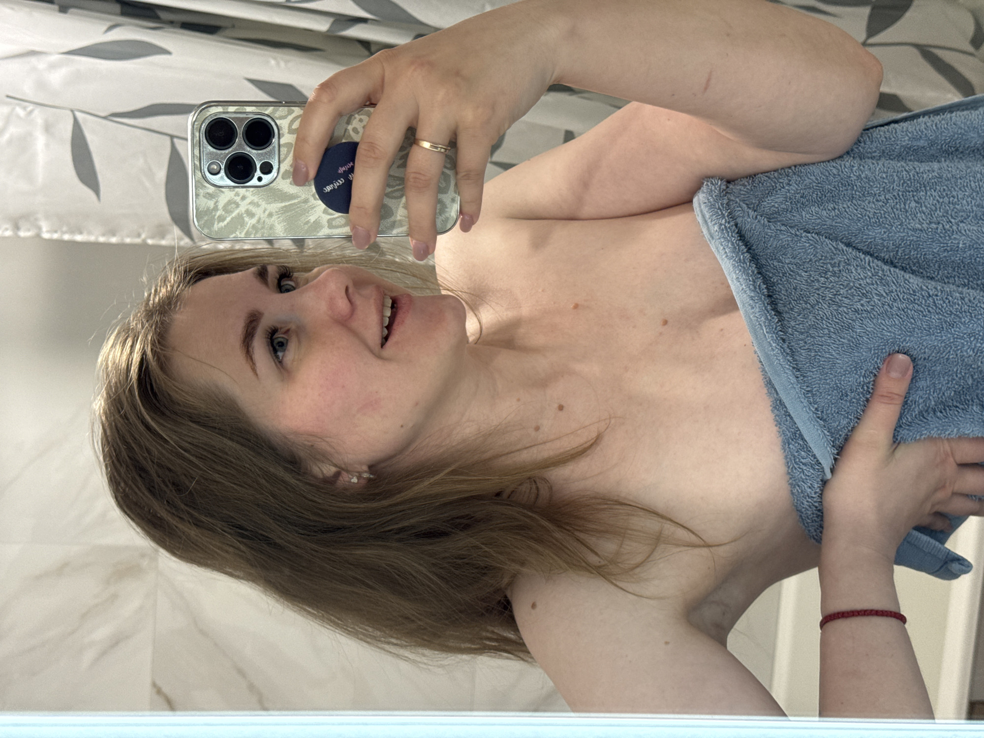 A selfie of a fair-skinned woman with blonde hair in a bathroom. She is holding a blue towel to her chest with her right hand, and her left hand holds an iPhone in a silver patterned case. The phone is positioned to capture her face in the mirror. The background shows a white shower curtain with grey leaf patterns and a white marble-like wall. A red bracelet is visible on her right wrist. There is no evidence suggesting the location is Ферма, Russia.